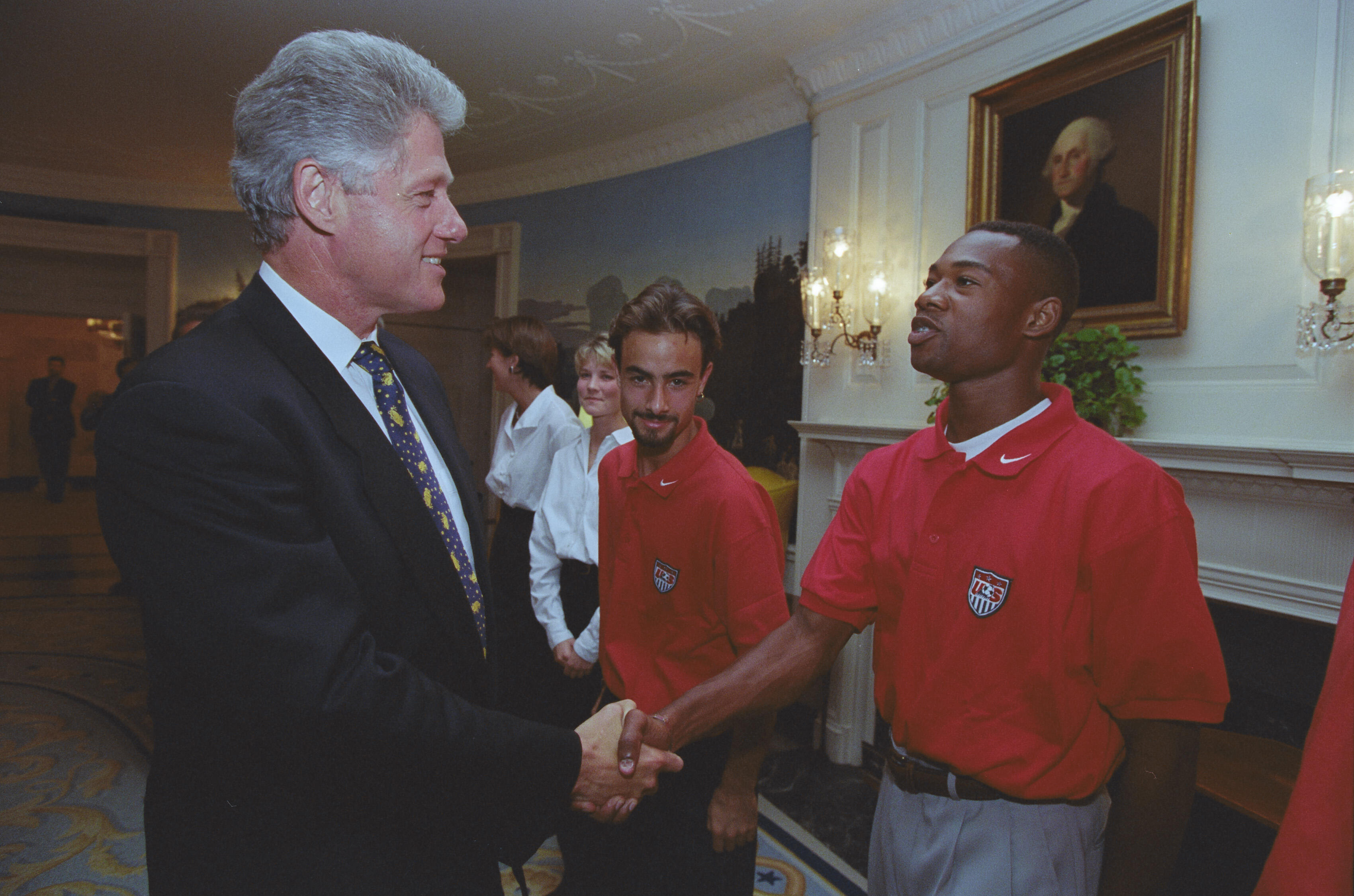 8A medium shot capturing President Bill Clinton on the left, smiling as he shakes hands with a Black soccer player wearing a red U.S. soccer team polo shirt. Another team member with a goatee stands directly behind the player, looking toward the camera. In the background, two women in white collared shirts stand near a scenic nature mural. The room features a white fireplace mantel, an oil portrait of George Washington, and ornate wall sconces.