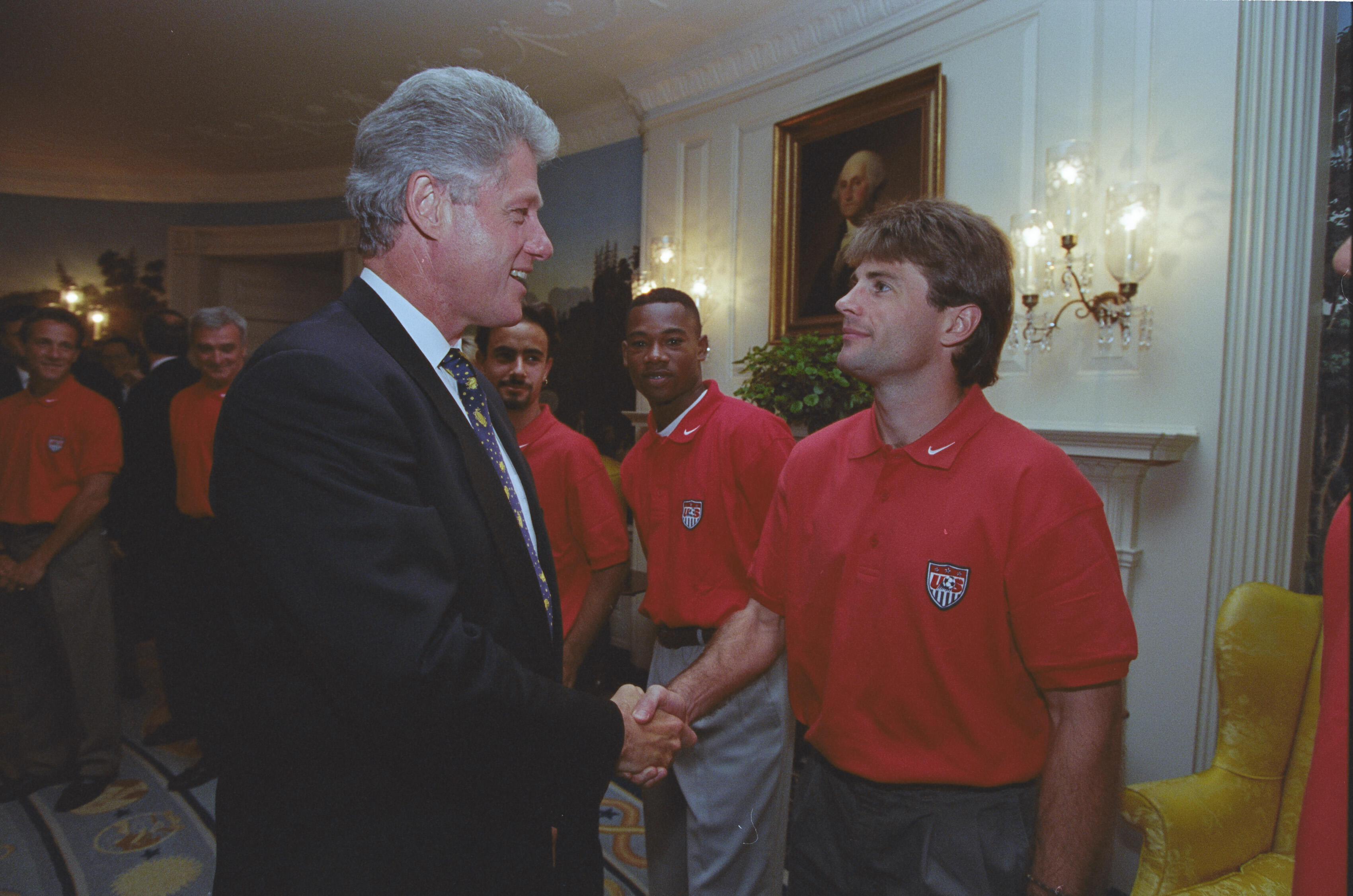 A photograph of President Bill Clinton shaking hands with a man wearing a red U.S. Soccer polo shirt inside the White House. Clinton, in profile on the left, wears a dark suit and patterned tie. The man looks directly at him as they shake hands. In the background, other members of the U.S. soccer team look on, and a framed portrait of George Washington hangs on the wall.