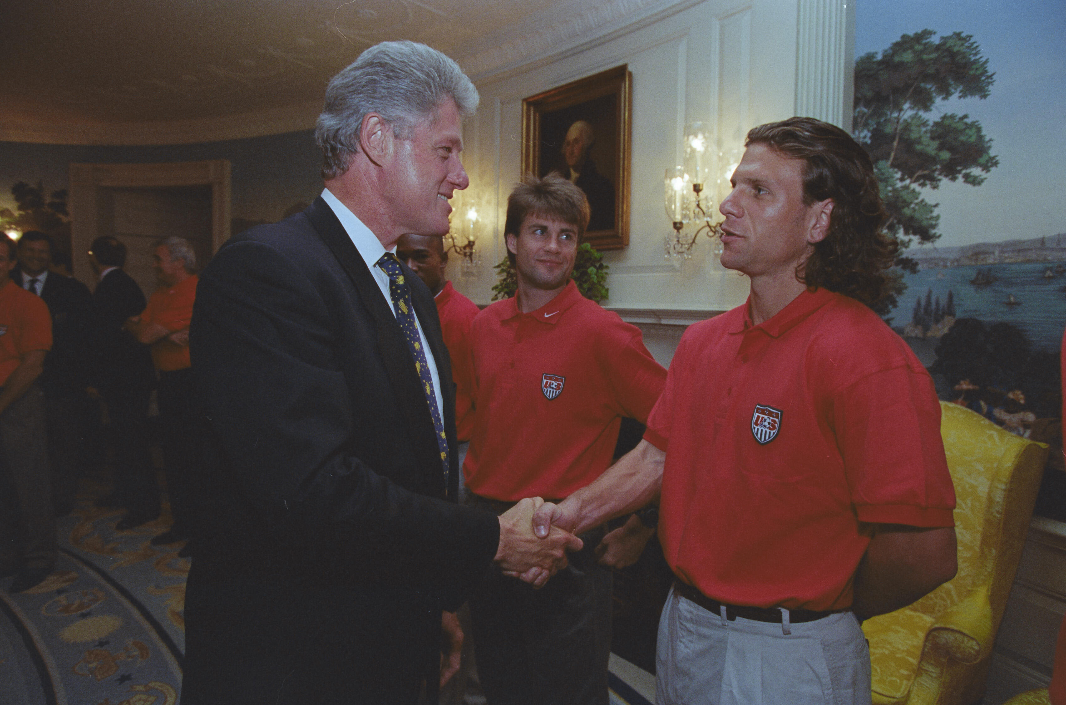 A medium shot capturing President Bill Clinton on the left, smiling as he shakes hands with a soccer player with long, wavy brown hair wearing a red U.S. soccer team polo shirt. Another team member with feathered brown hair stands directly between them, looking toward the pair. The setting features a white wall with a framed portrait of George Washington, ornate lit wall sconces, a scenic nature mural on the right, and a yellow upholstered chair.