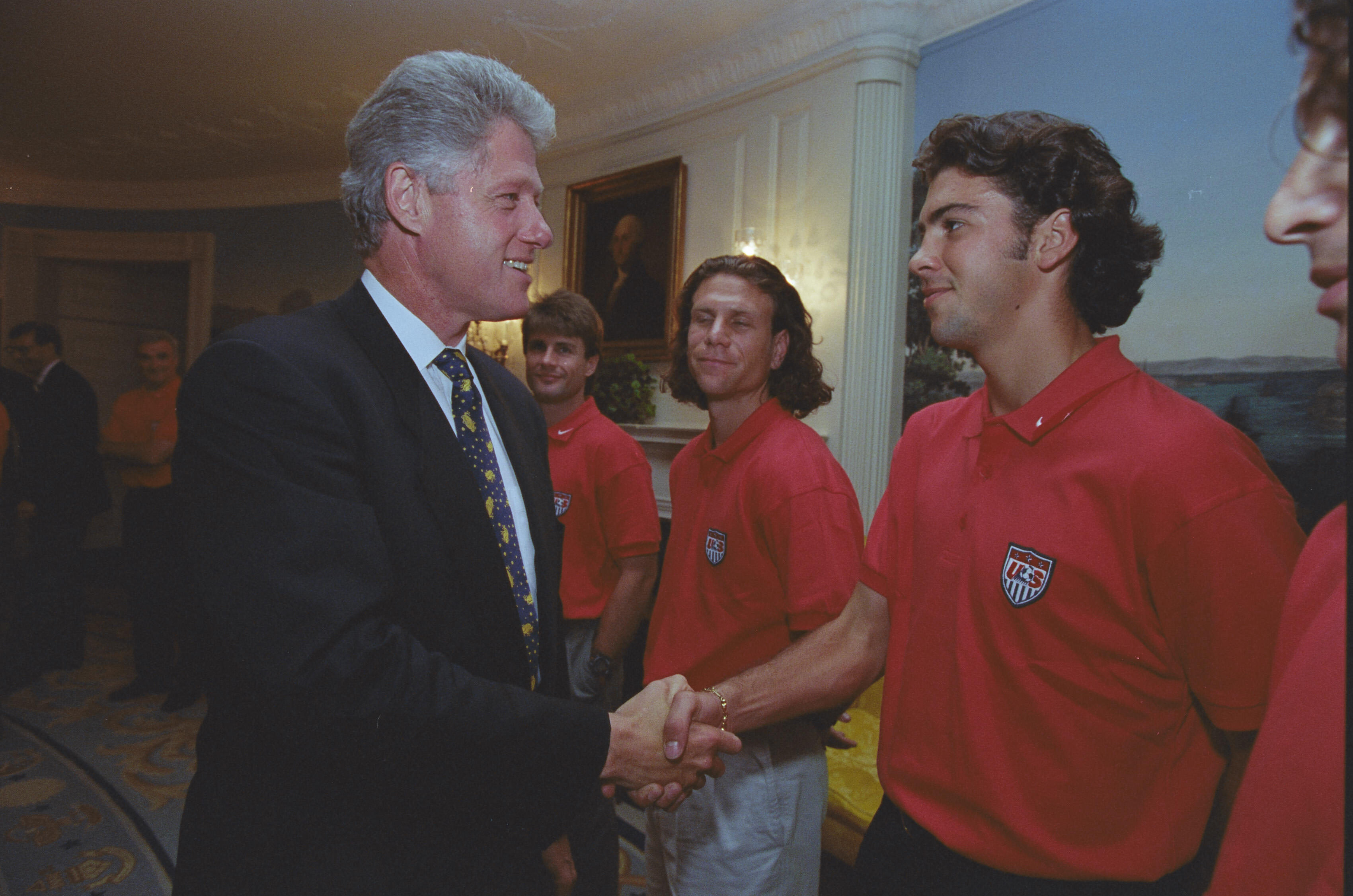 President Bill Clinton shaking hands with US Men's National Soccer Team player inside the White House. Clinton, shown in profile on the left wearing a dark suit and a blue patterned tie, smiles warmly. He standing on the right wearing a red U.S. Soccer polo shirt, looks toward the President. In the background between them, teammates watch the interaction, with a framed portrait of George Washington visible on the wall behind them.