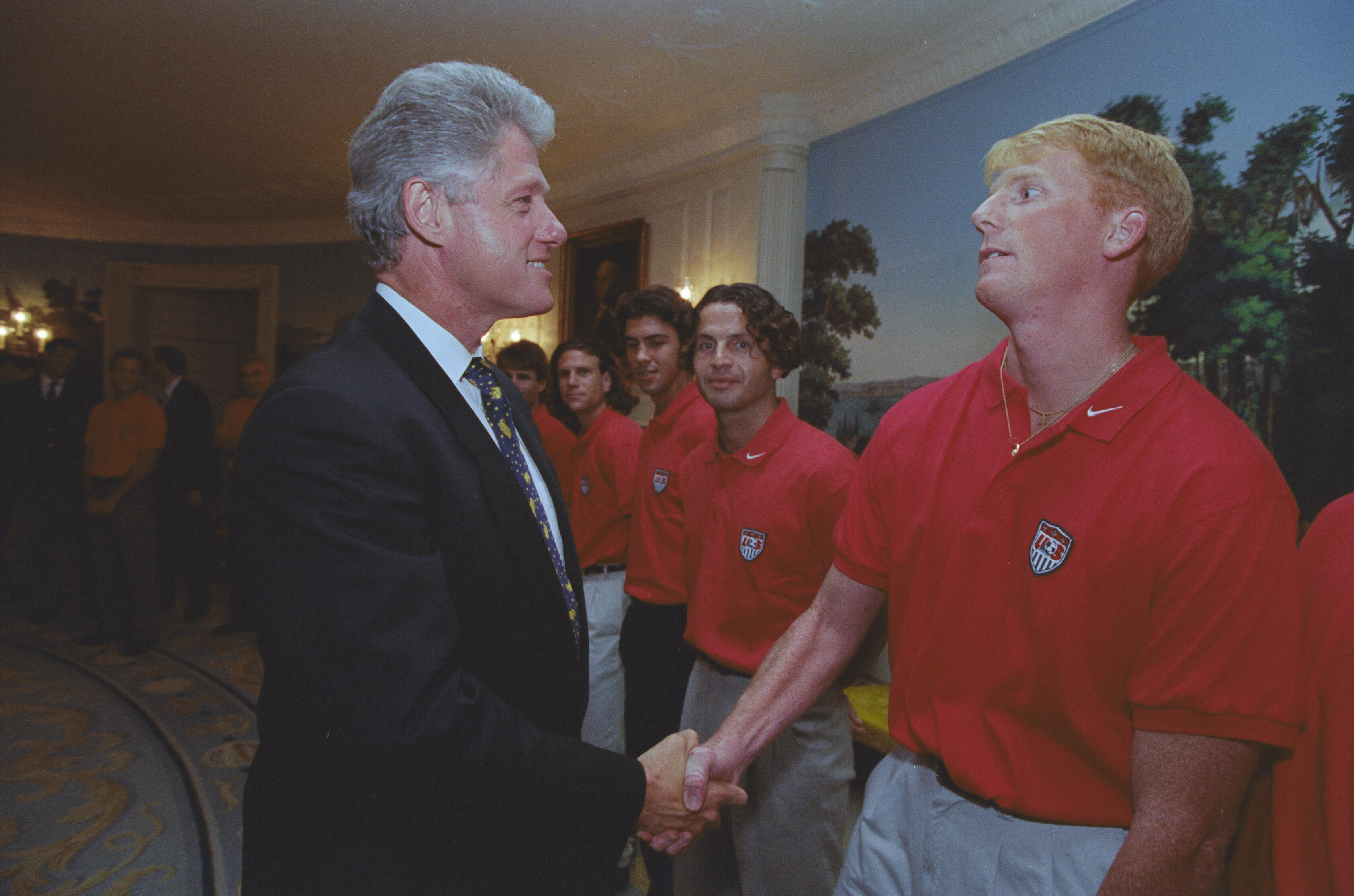 President Bill Clinton shakes hands with a US soccer player wearing a red team polo shirt in the White House. Several other players stand in line behind him.