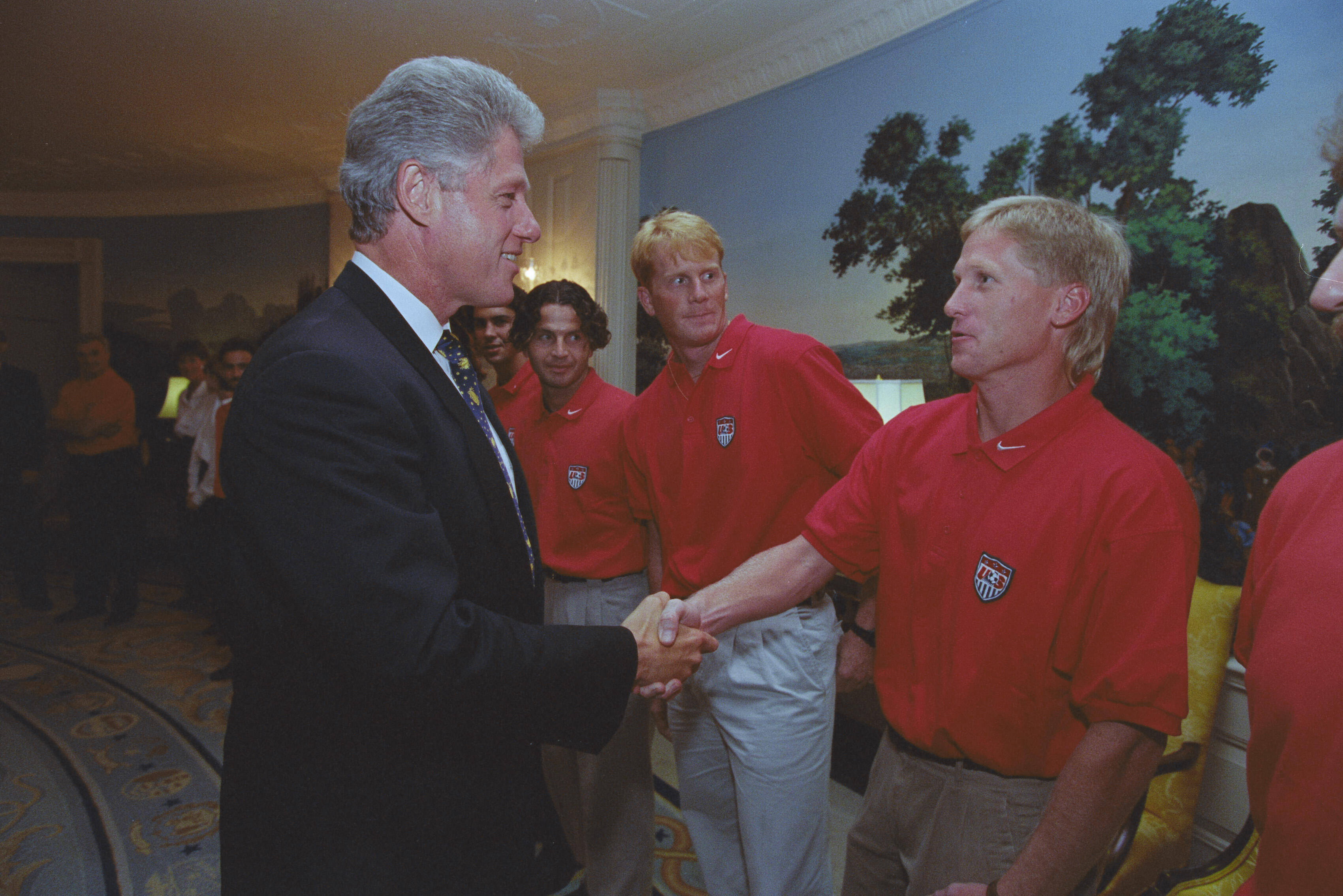 President Bill Clinton shakes hands with a blonde US soccer player in a red team polo shirt inside the White House. The player previously shaking hands stands behind him, along with other teammates.