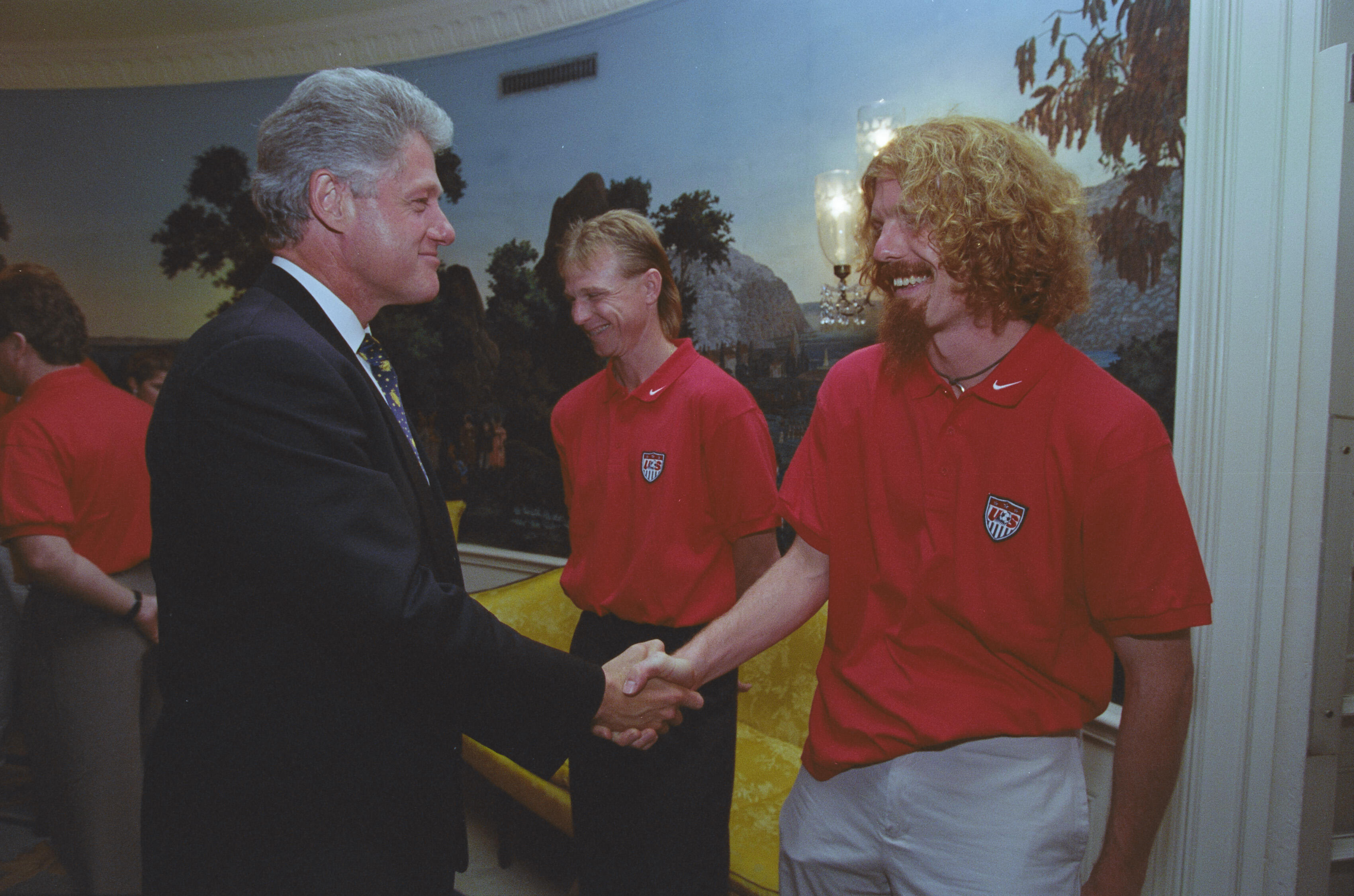 A medium shot capturing President Bill Clinton on the left, smiling as he shakes hands with a soccer player featuring long, curly blonde hair and a goatee. The player wears a red U.S. soccer team polo shirt. In the background between them, another teammate with lighter hair looks down and smiles. The room features a scenic nature mural on the wall, an ornate light fixture, and a bright yellow upholstered sofa behind the men.