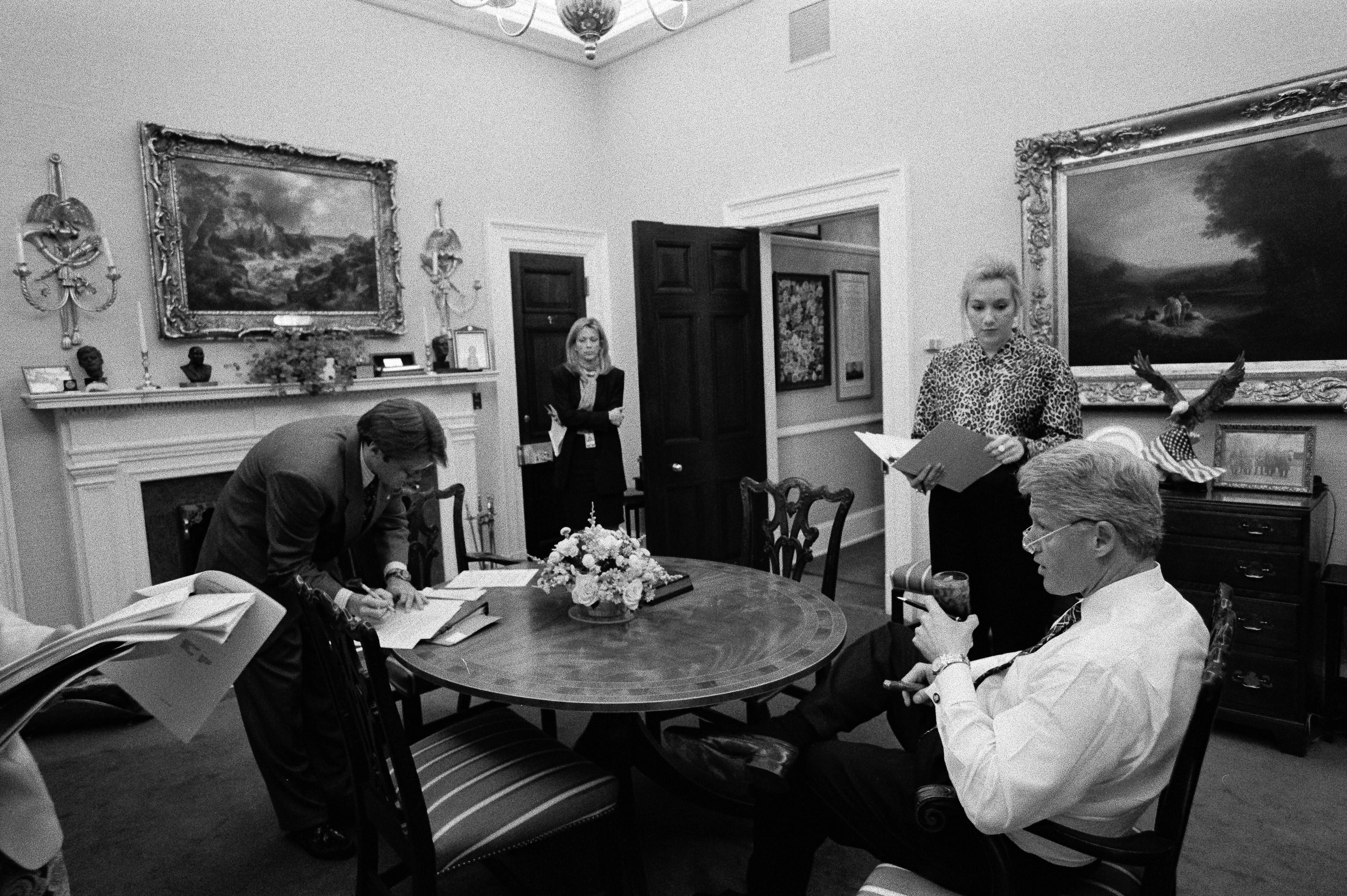 Black and white photograph. President Clinton meets with Stephanie Streett, Stephen Goodin and Nancy Hernreich in the Oval Office Dining Room. Heinreich and Streett stand while the President sits drinking a soft drink. Goodin stands leaning over the dining table.  