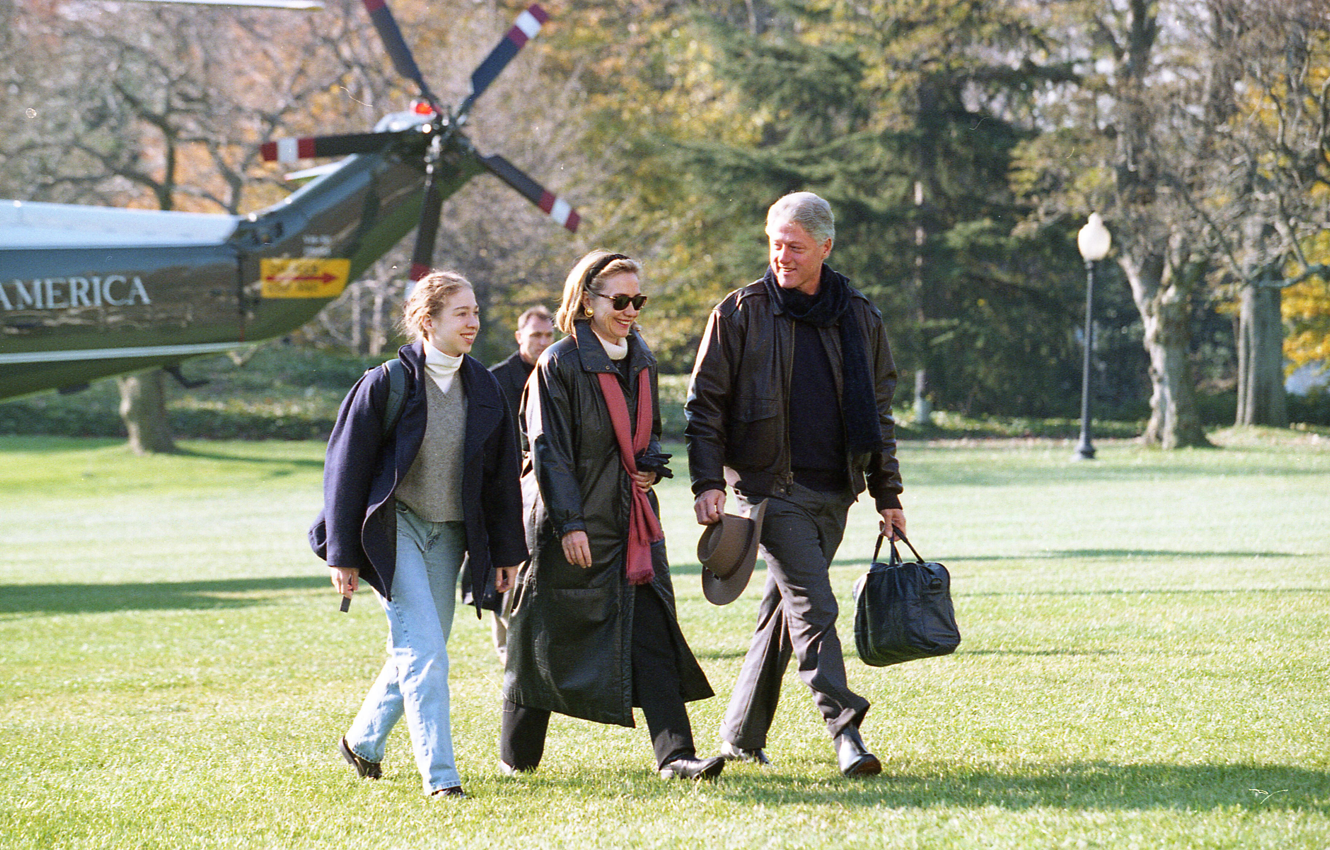 President Clinton, Hillary Rodham Clinton, and Chelsea Clinton walk across the White House South Lawn together. President Clinton caries a bag in his left hand and a hat in his right hand. The Marine Helicopter, Marine 1, can be seen in the background.