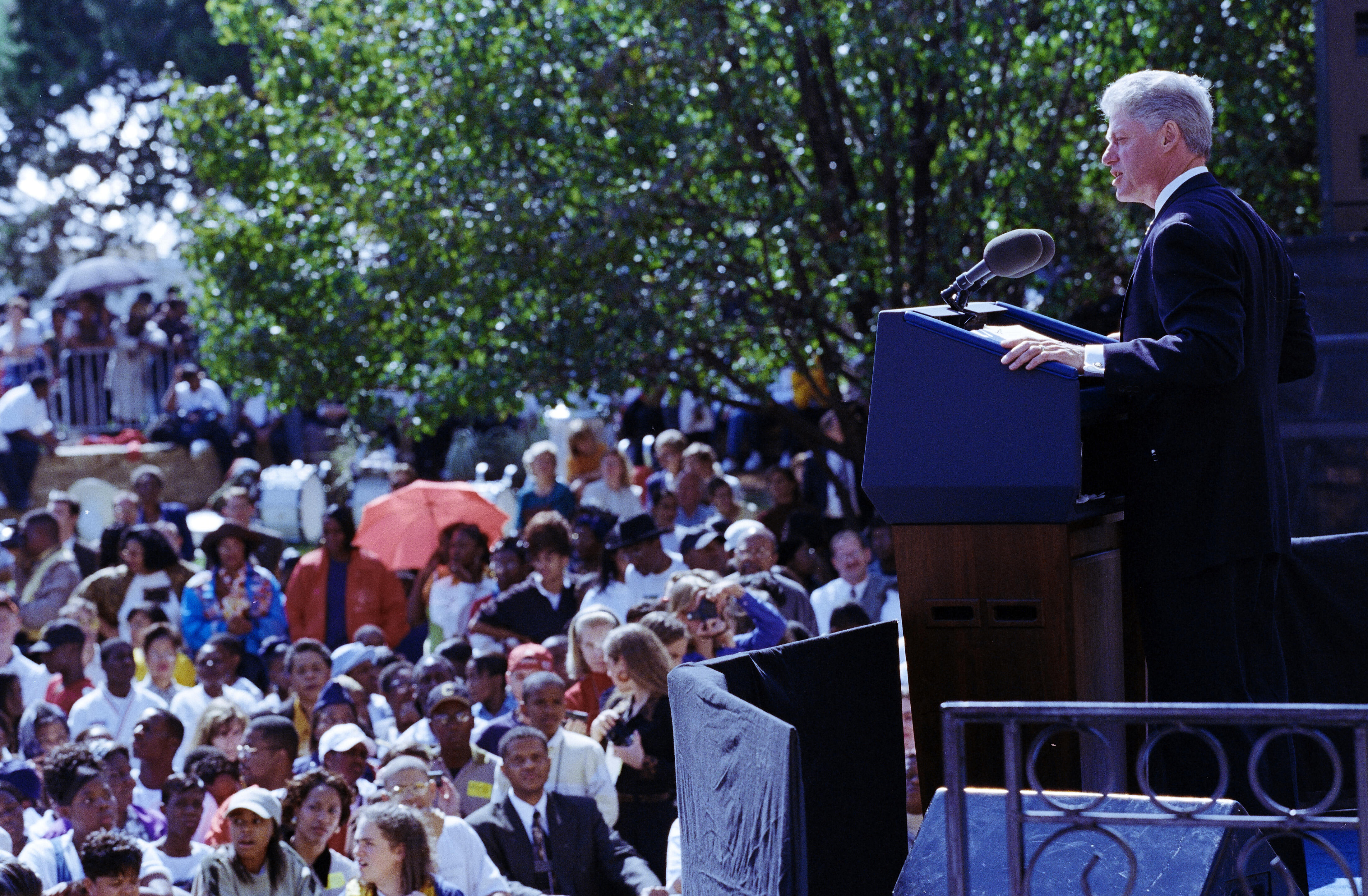 President Clinton stands on a stage, behind a podium, looking out toward a crowd of people.