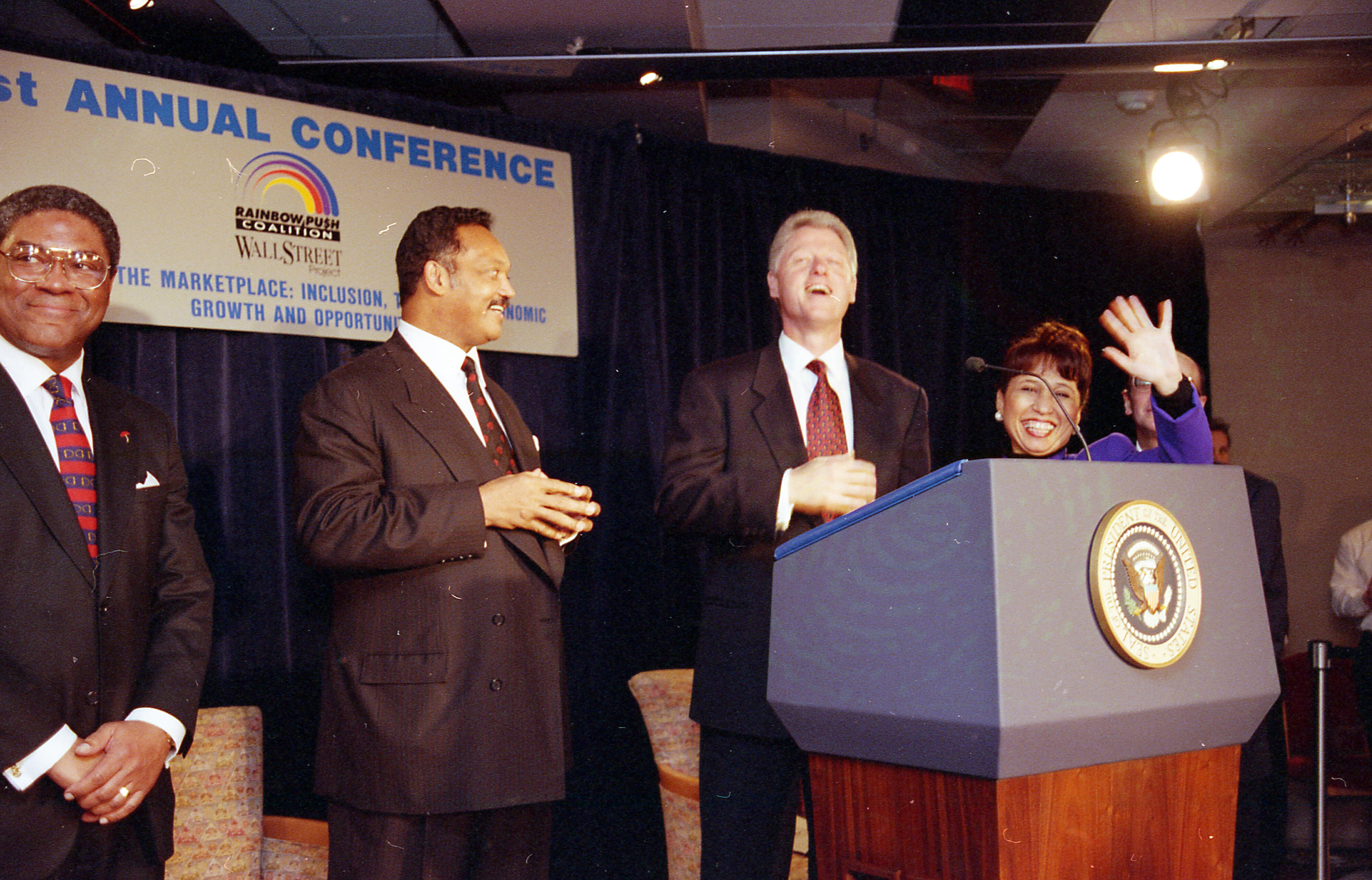 President Clinton laughs with Rev. Jesse Jackson, Sec. Alexis Herman. They all stand behind a podium.