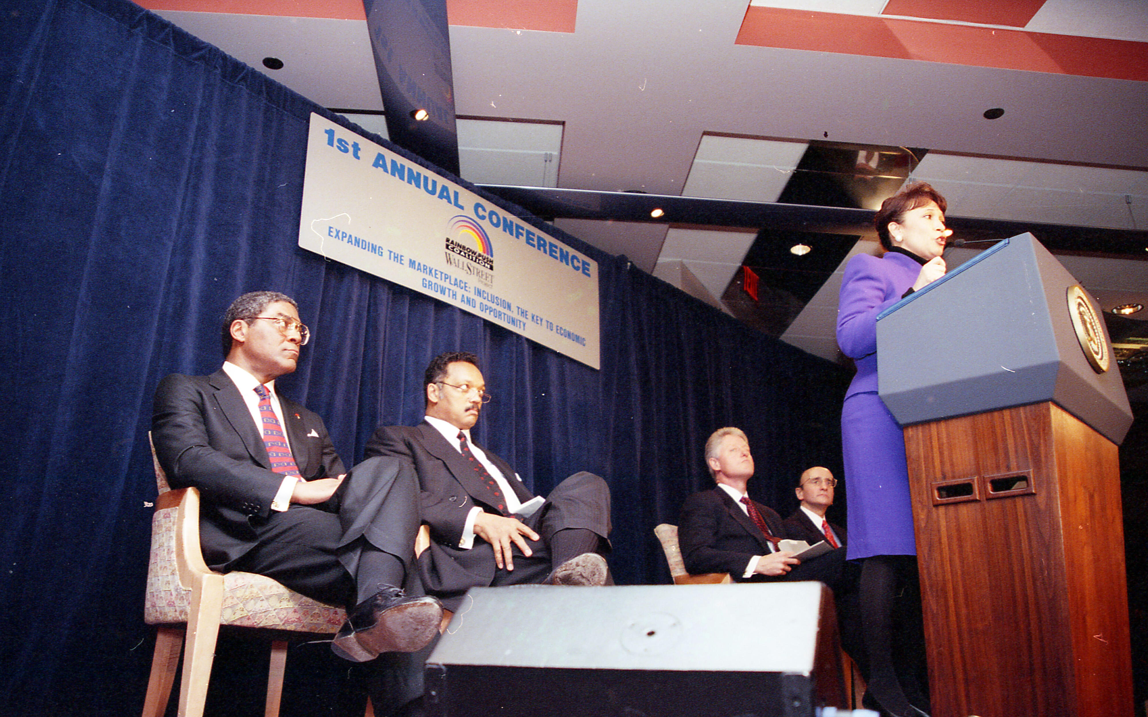 Sec. Alexis Herman at the podium a. Seated behind Herman are, President Clinton; Rev. Jesse Jackson; and Chairman of the New York Stock Exchange, Richard Grasso. 