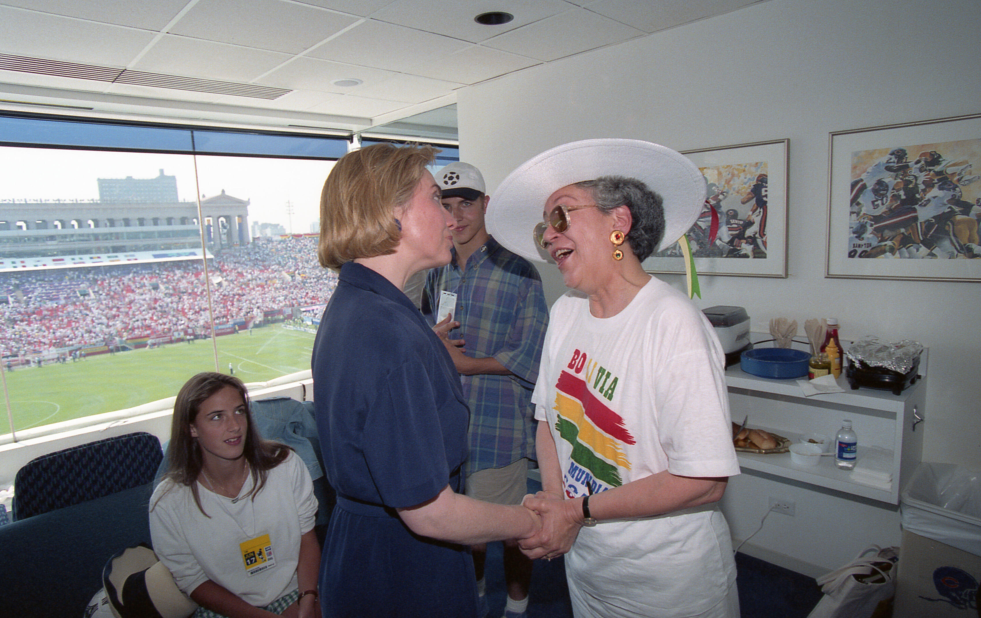 In the foreground, Hillary Rodham Clinton (left), then-First Lady of the United States, stands in a dark blue dress while shaking hands and speaking with Ximena Iturralde (right), then-First Lady of Bolivia. Iturralde wears a white t-shirt featuring the word "BOLIVIA" and a colorful map above the WorldCupUSA94 logo, topped by a wide-brimmed white hat.