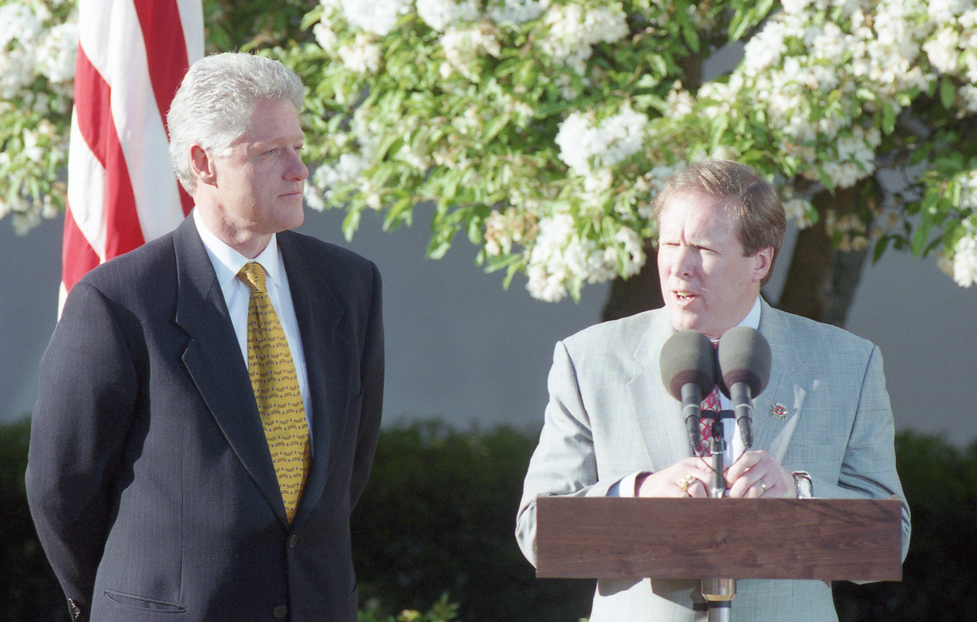 An eye-level, medium shot shows Bill Clinton, left, and President Kevin Payne , right, standing outdoors against a backdrop of blooming white trees. Clinton, wearing a navy blue suit and a patterned gold tie, stands with his hands behind his back, looking toward the man at the podium. The man at the podium, in a light gray suit and a red patterned tie, speaks into two microphones. An American flag is partially visible in the upper left corner.