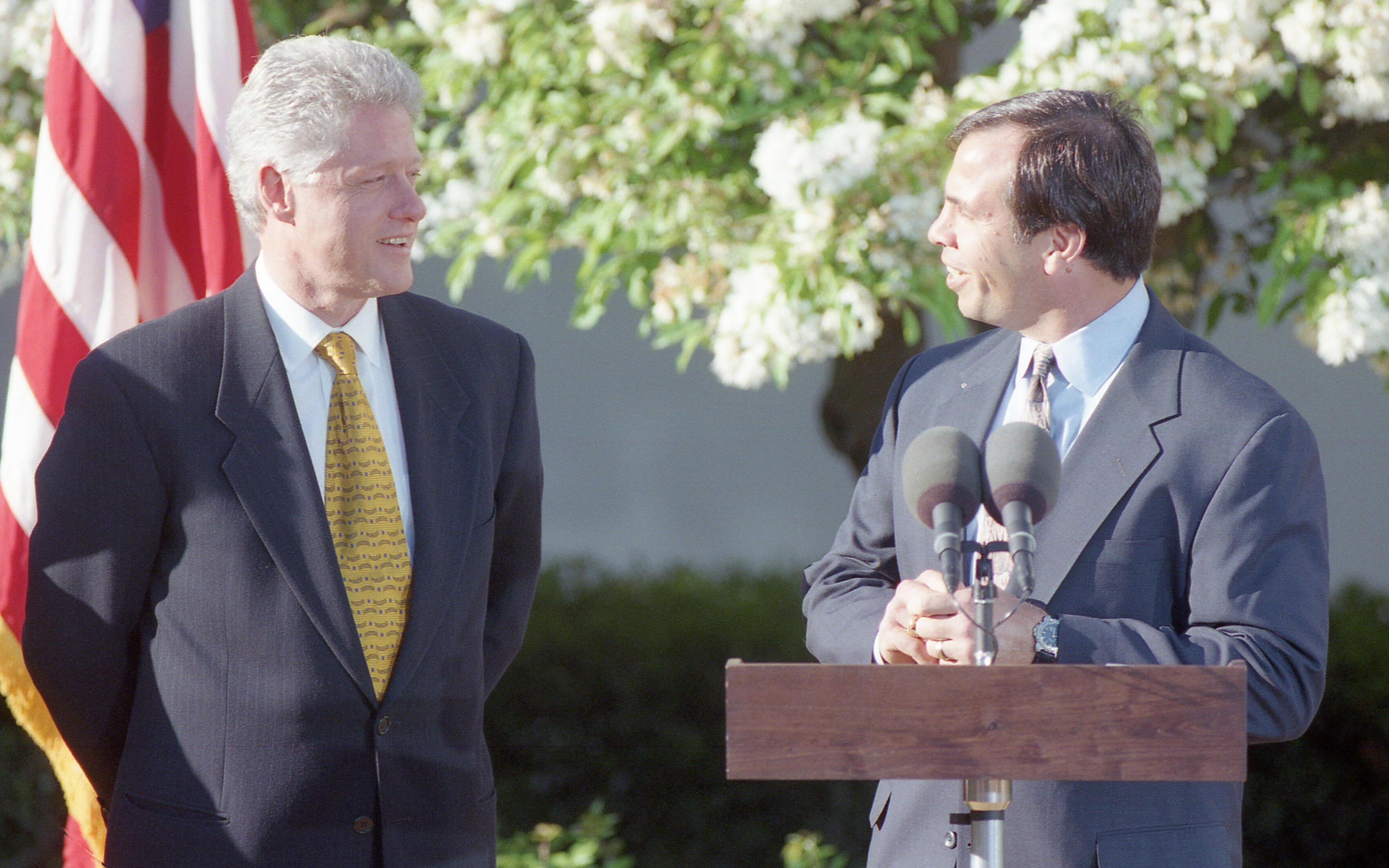 An eye-level, medium shot shows Bill Clinton, left, and Coach Bruce Arena, right, standing outdoors against a backdrop of blooming white trees. Clinton, wearing a navy blue suit and a patterned gold tie, stands with his hands behind his back, looking toward the man at the podium with a smile. The man at the podium, in a dark navy suit and a patterned silver tie, also smiles and speaks into two microphones. An American flag is partially visible in the upper left corner.