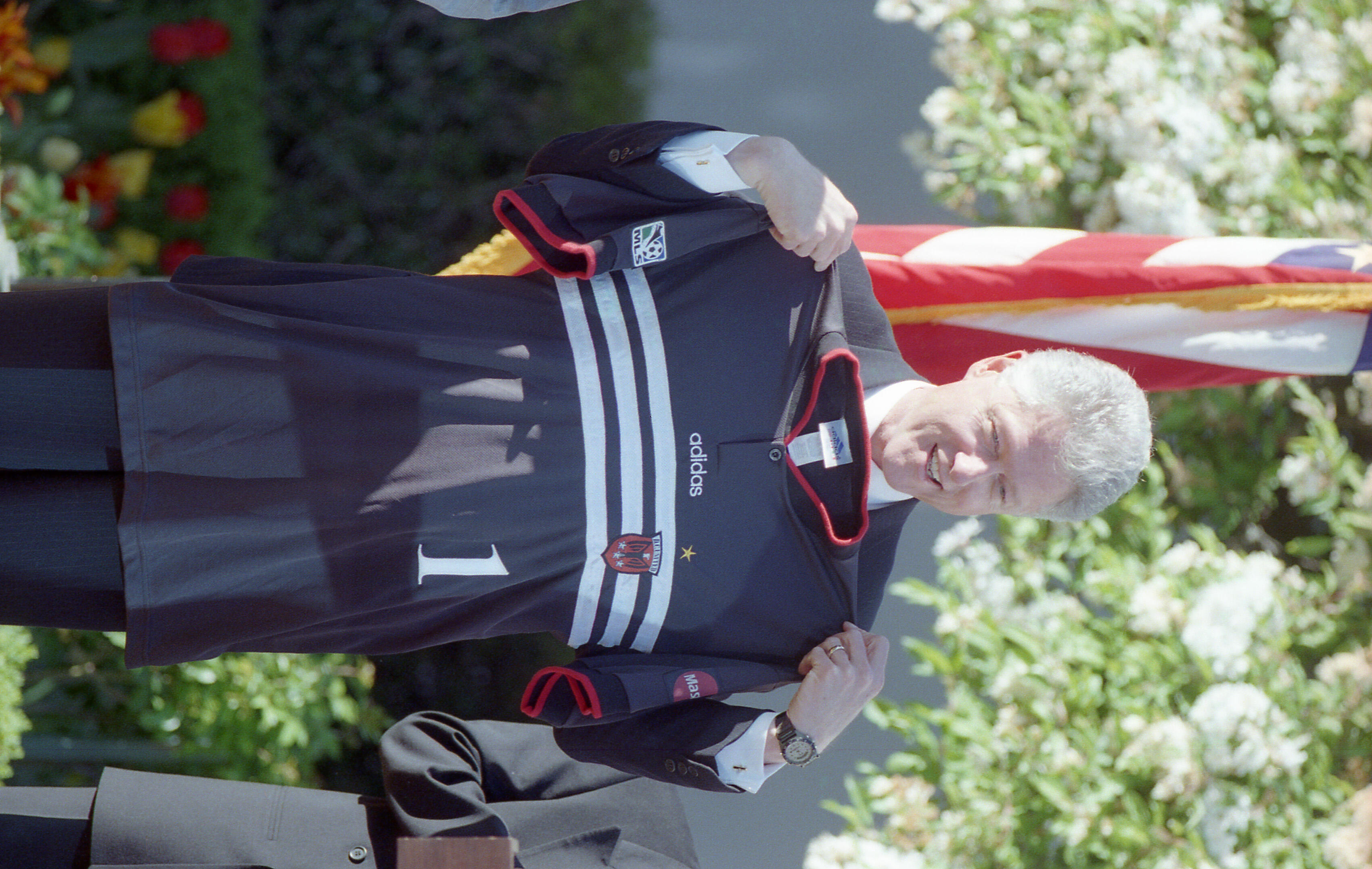 This medium, eye-level shot captures Bill Clinton smiling as he holds up a black D.C. United soccer jersey. He is dressed in a dark suit and stands in front of a blooming white tree, with a portion of the American flag visible in the background. The 1996-98 home jersey features white horizontal stripes across the chest, the Adidas logo, and the number "1". A small red and white D.C. United crest is visible on the left side.