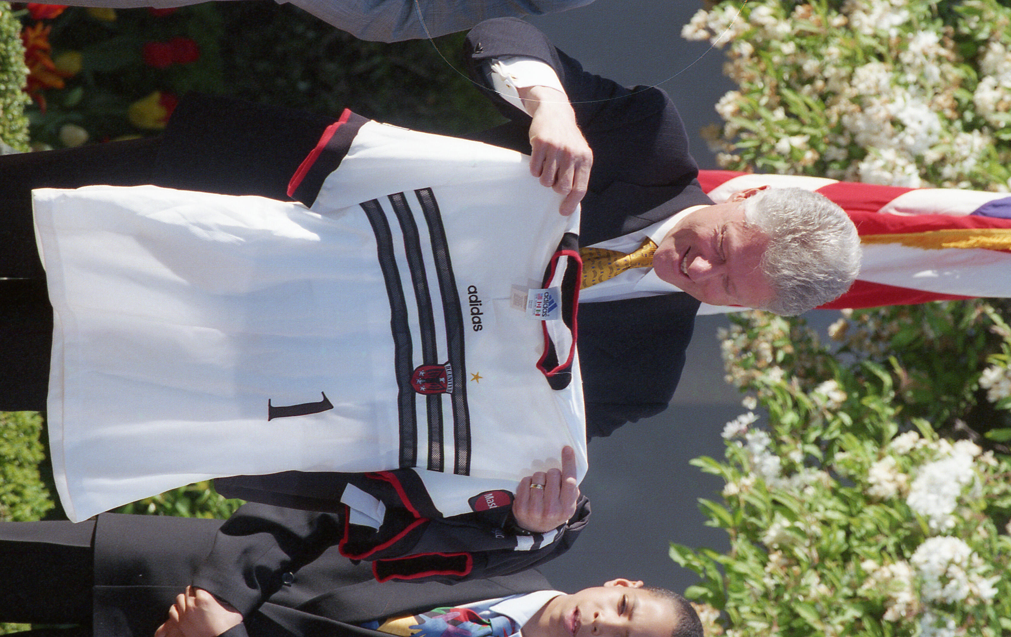 An eye-level, medium shot shows Bill Clinton smiling as he holds up the front of a white 1996-98 D.C. United away jersey. He is dressed in a dark suit and patterned gold tie, standing outdoors in front of a blooming white tree and a portion of the American flag.The jersey features three black horizontal stripes across the chest, the Adidas logo, and the number "1". A small red and white D.C. United crest is visible on the left side. To Clinton's right, a young boy in a dark suit and colorful tie looks on.