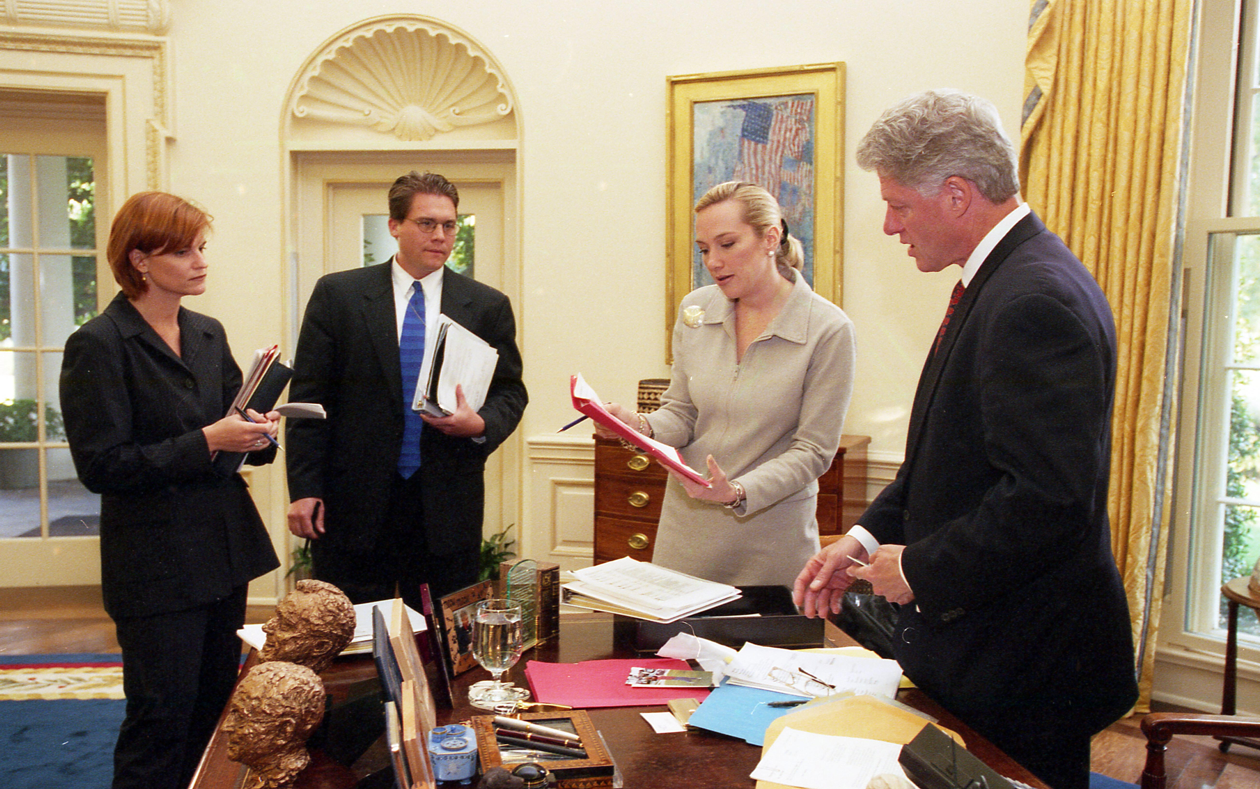 Color photograph. President Clinton meets with Stephanie Streett, Jennifer Palmieri, and Jeff Forbes in the White House Oval Office. All individuals stand near the President's desk. Streett is holding a document in front of President Clinton.