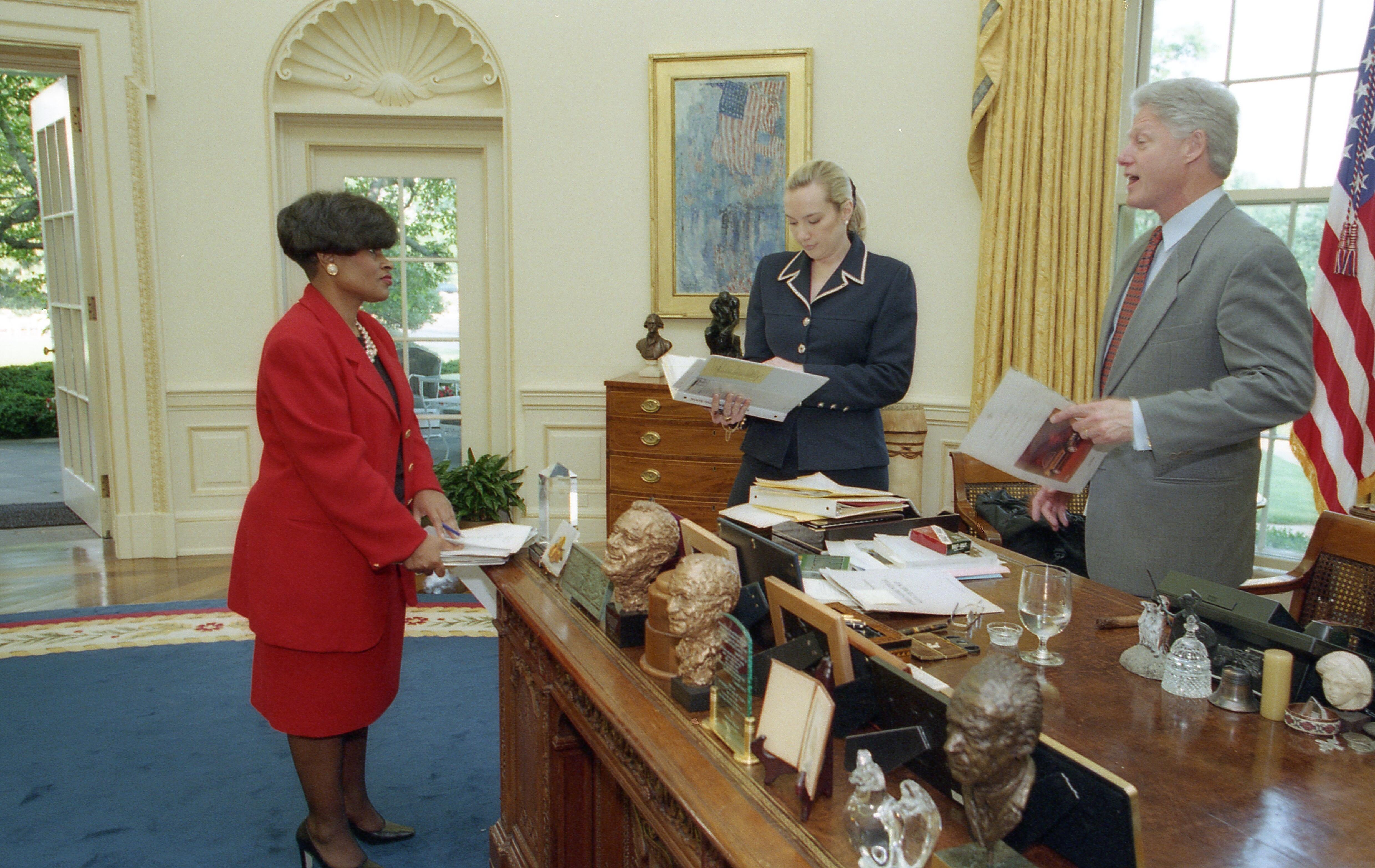 President Clinton, Stephanie Streett, and Minyon Moore work while standing around the Resolute Desk in the Oval Office.