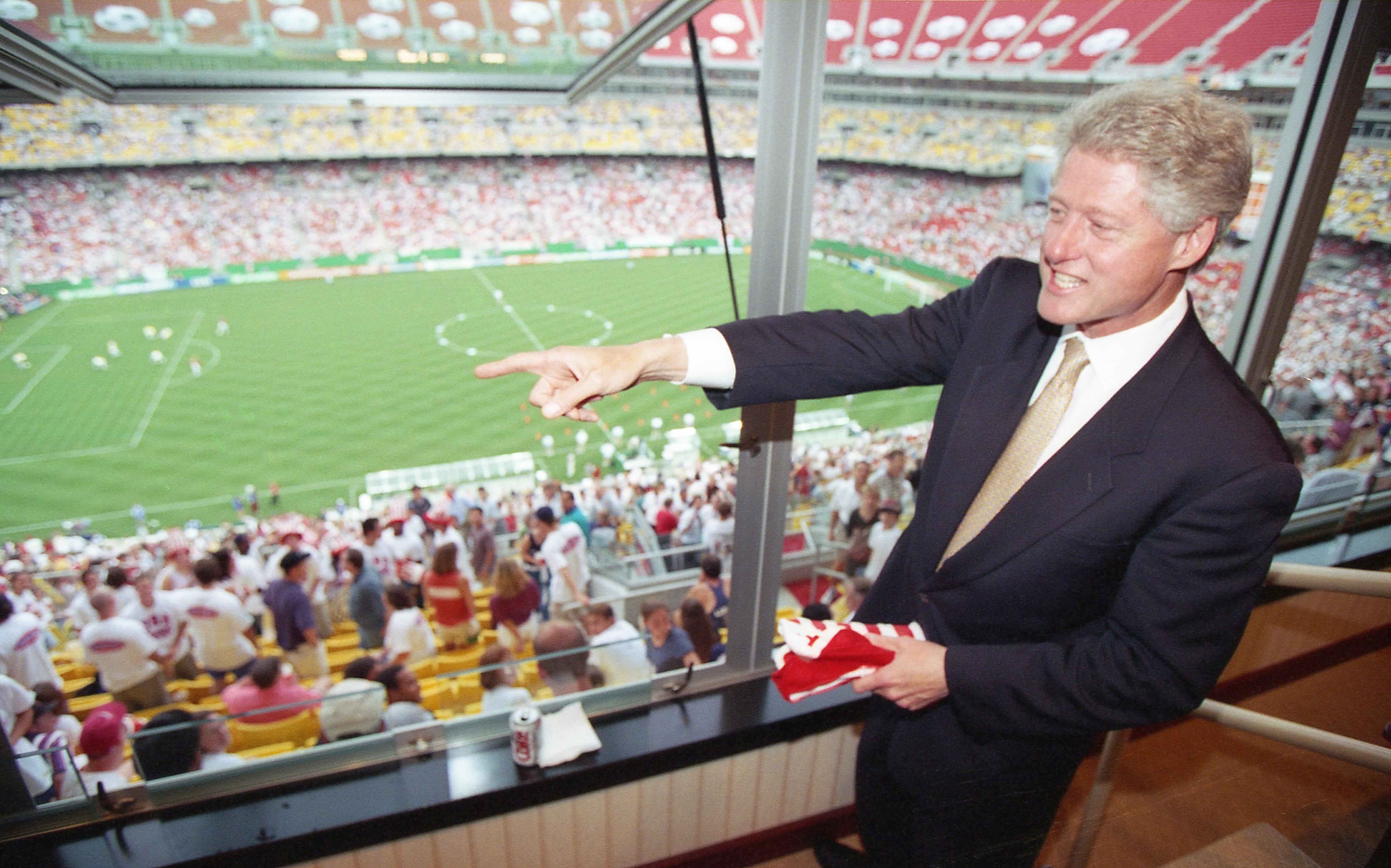 A photograph captures US President Bill Clinton looking out from a glass-walled stadium luxury box over a packed soccer stadium during a match. He wears a dark suit, white shirt, and gold tie, smiling as he points with his right hand toward the green field below. In his left hand, he holds a folded red and white garment. Below the luxury box, rows of spectators fill the yellow stadium seats, and a blurred soccer pitch is visible through the window.
