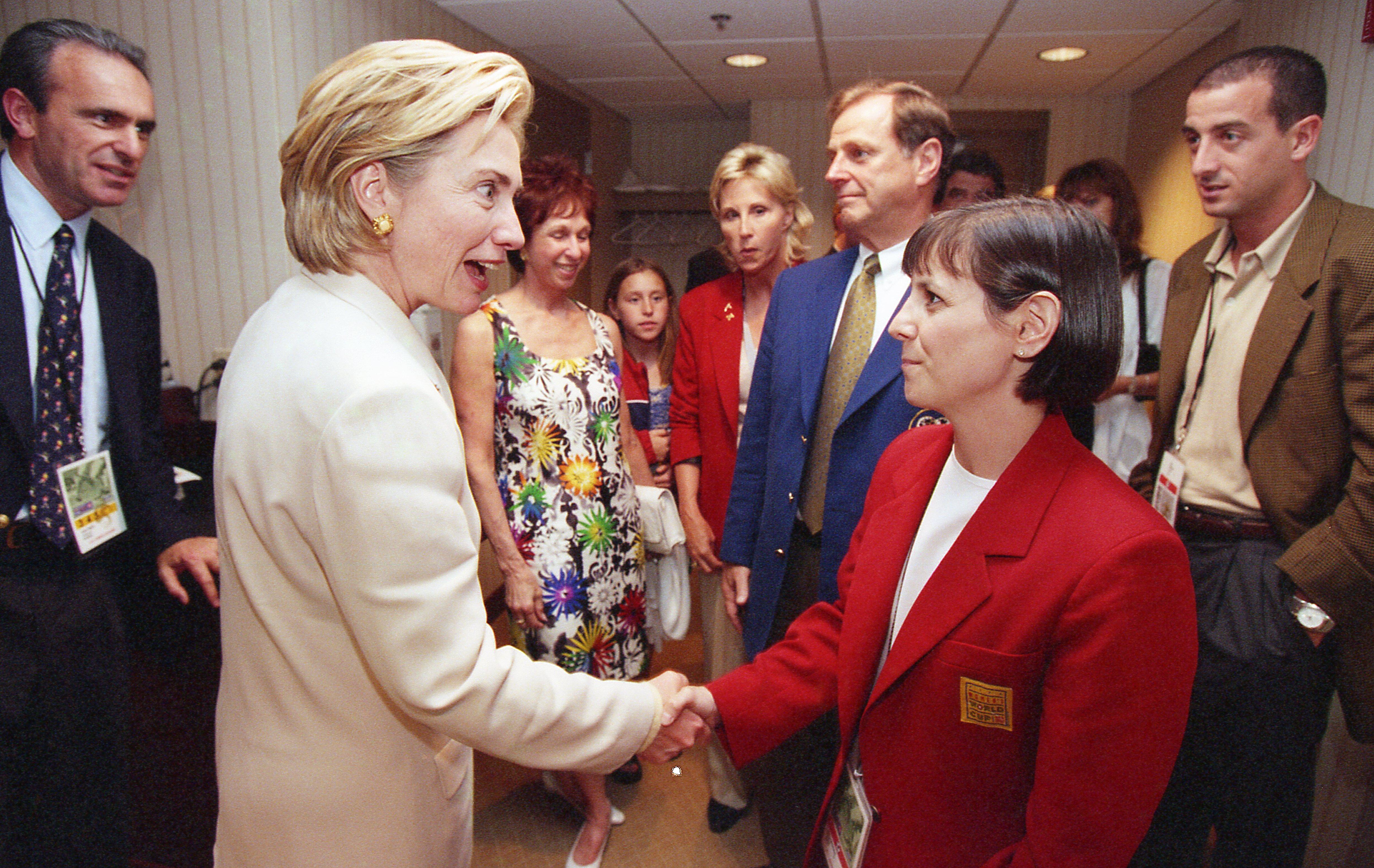 A photograph shows First Lady Hillary Clinton shaking hands with an official inside a stadium corridor. Hillary Clinton, positioned on the left in profile, wears a cream-colored pantsuit and smiles while greeting a woman with short dark hair wearing a bright red blazer with a gold 1999 FIFA Women's World Cup patch. Several onlookers stand in the background, including a man in a dark suit and patterned tie on the far left, a woman in a colorful floral dress, a man in a blue sport coat and gold tie, and a man
