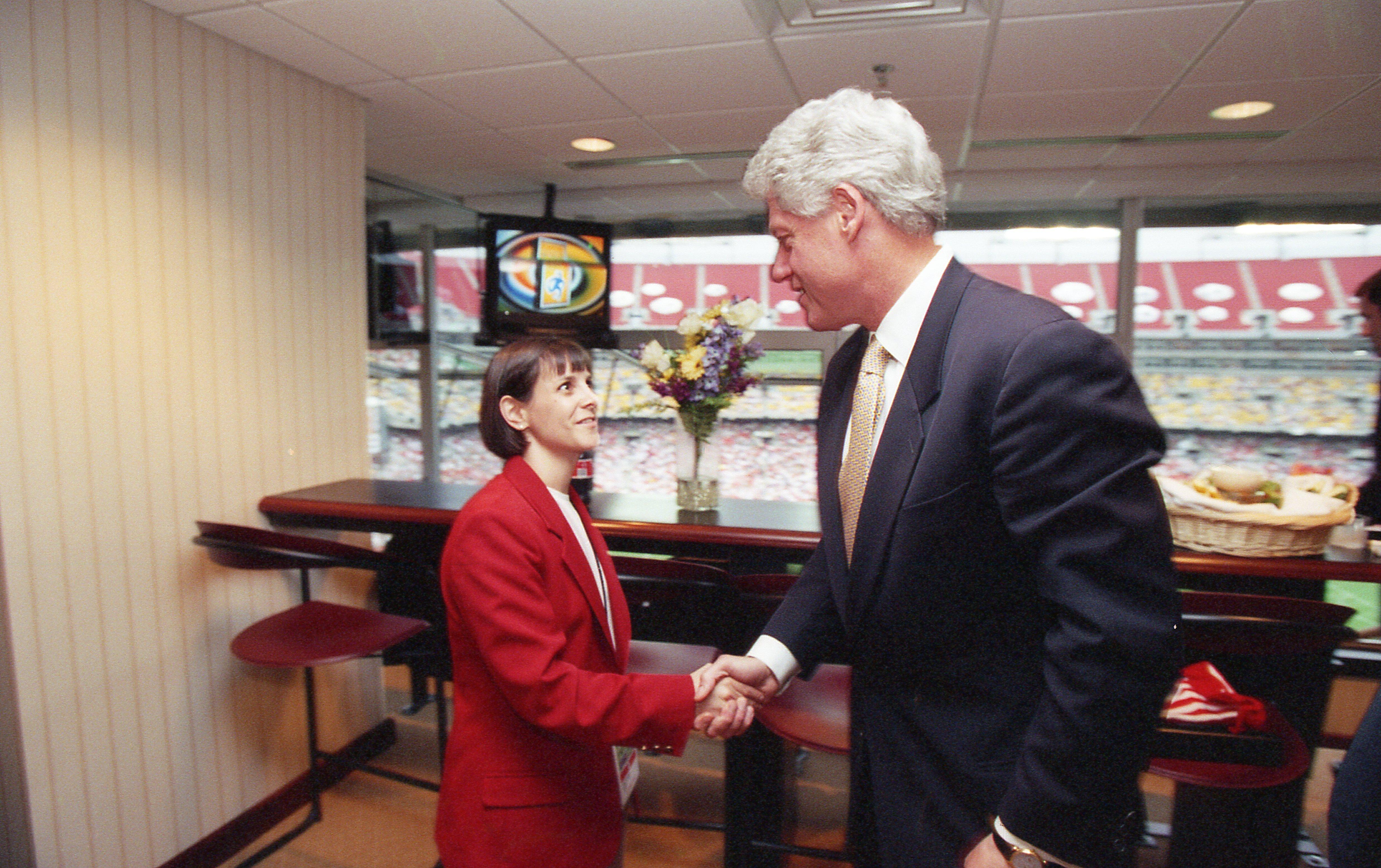 A photograph captures Marla Messing, President and CEO of the 1999 FIFA Women's World Cup Organizing Committee, shaking hands with US President Bill Clinton inside a stadium luxury box. On the left, Messing wears a bright red tournament official blazer over a white shirt as she looks up and smiles. On the right, Clinton stands in profile wearing a dark suit and gold tie, smiling down as he shakes her hand. In the background, a glass window overlooks rows of stadium seating, with a bar counter, stools, and a