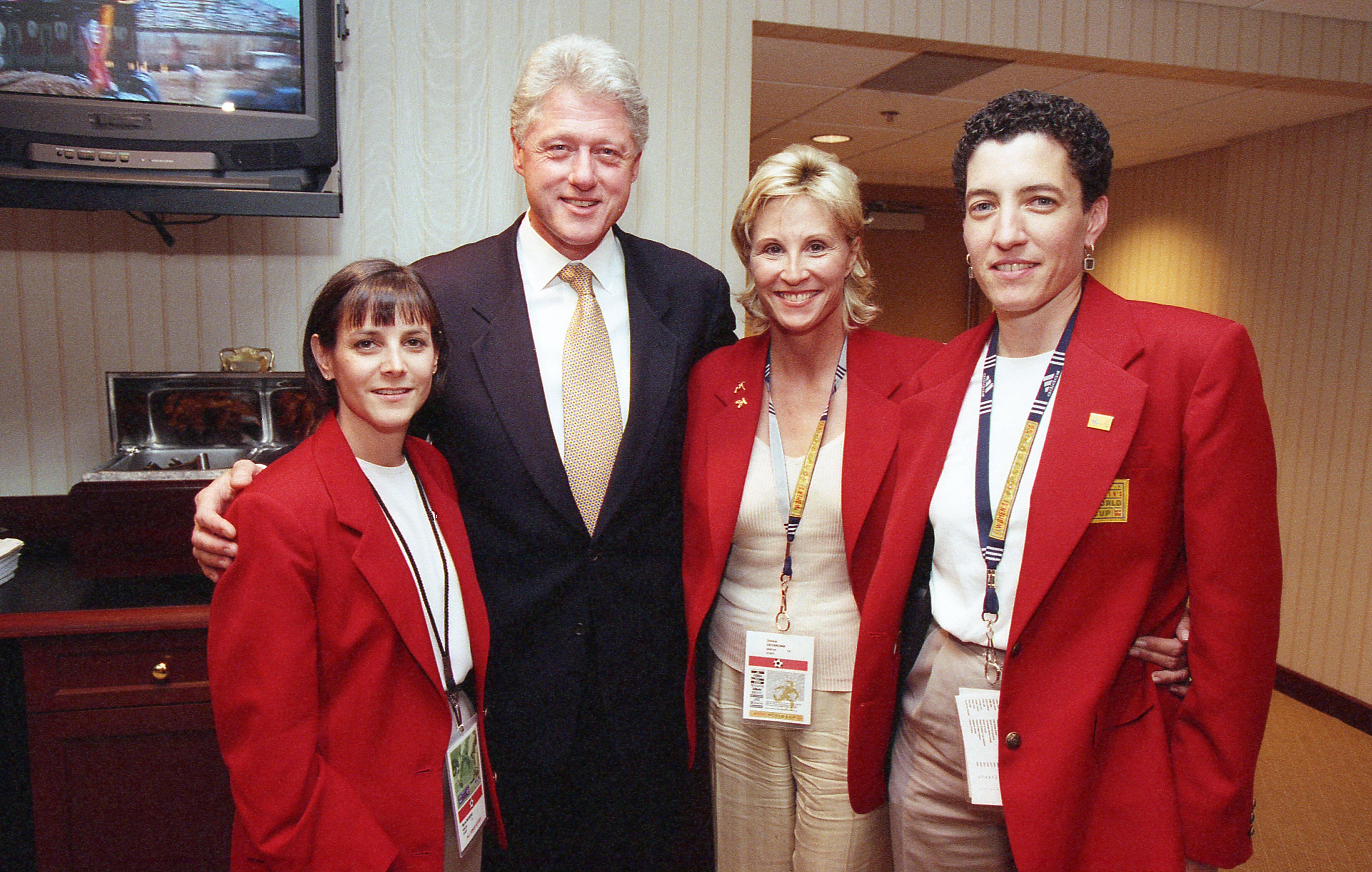 A photograph shows Bill Clinton posing with three women wearing matching red blazers and credentials around their necks, standing indoors with a television and buffet setup in the background.