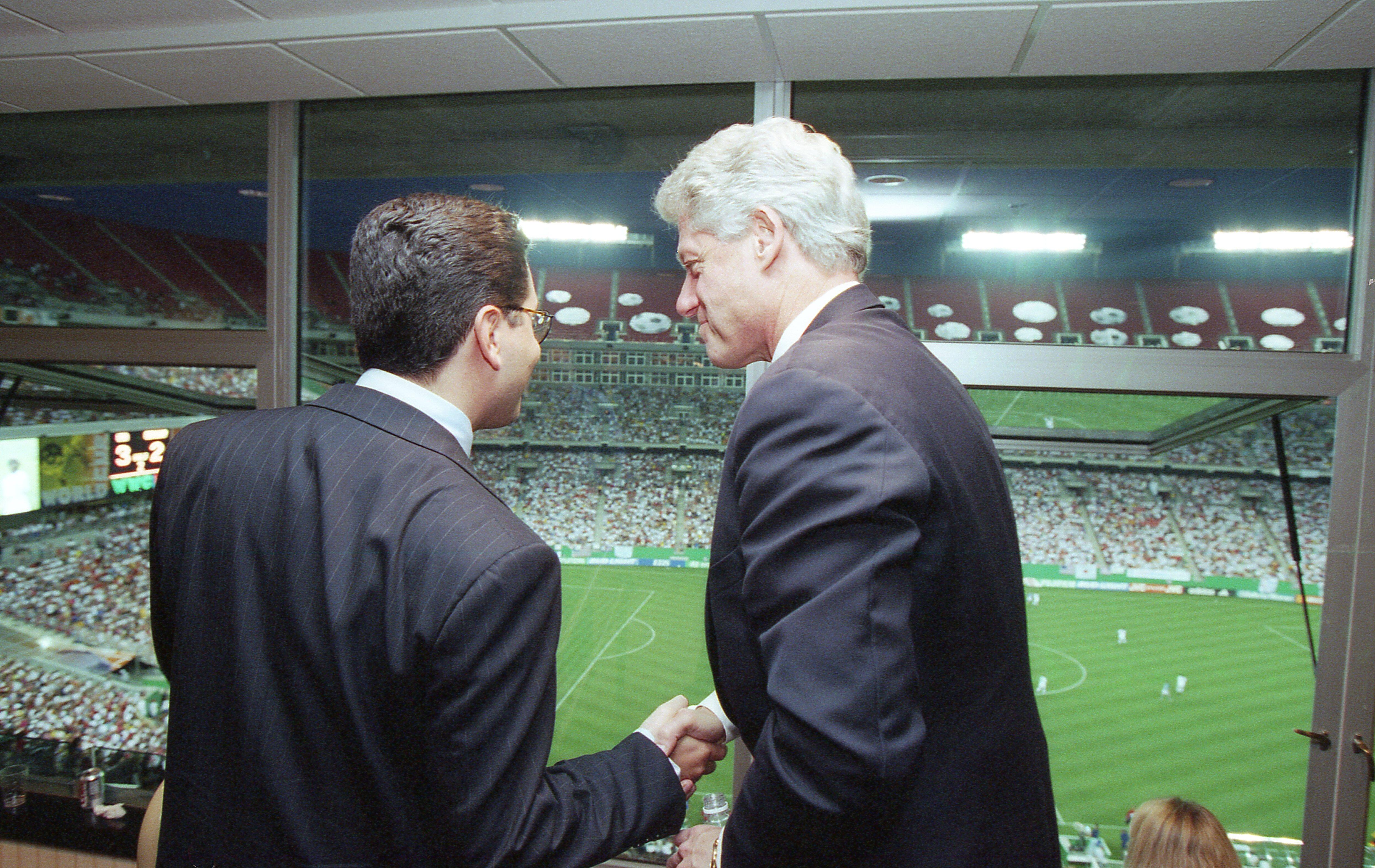 A photograph shows Bill Clinton shaking hands with a man wearing a pinstripe suit, viewed from behind as they stand inside a stadium suite looking out over a soccer field filled with spectators.