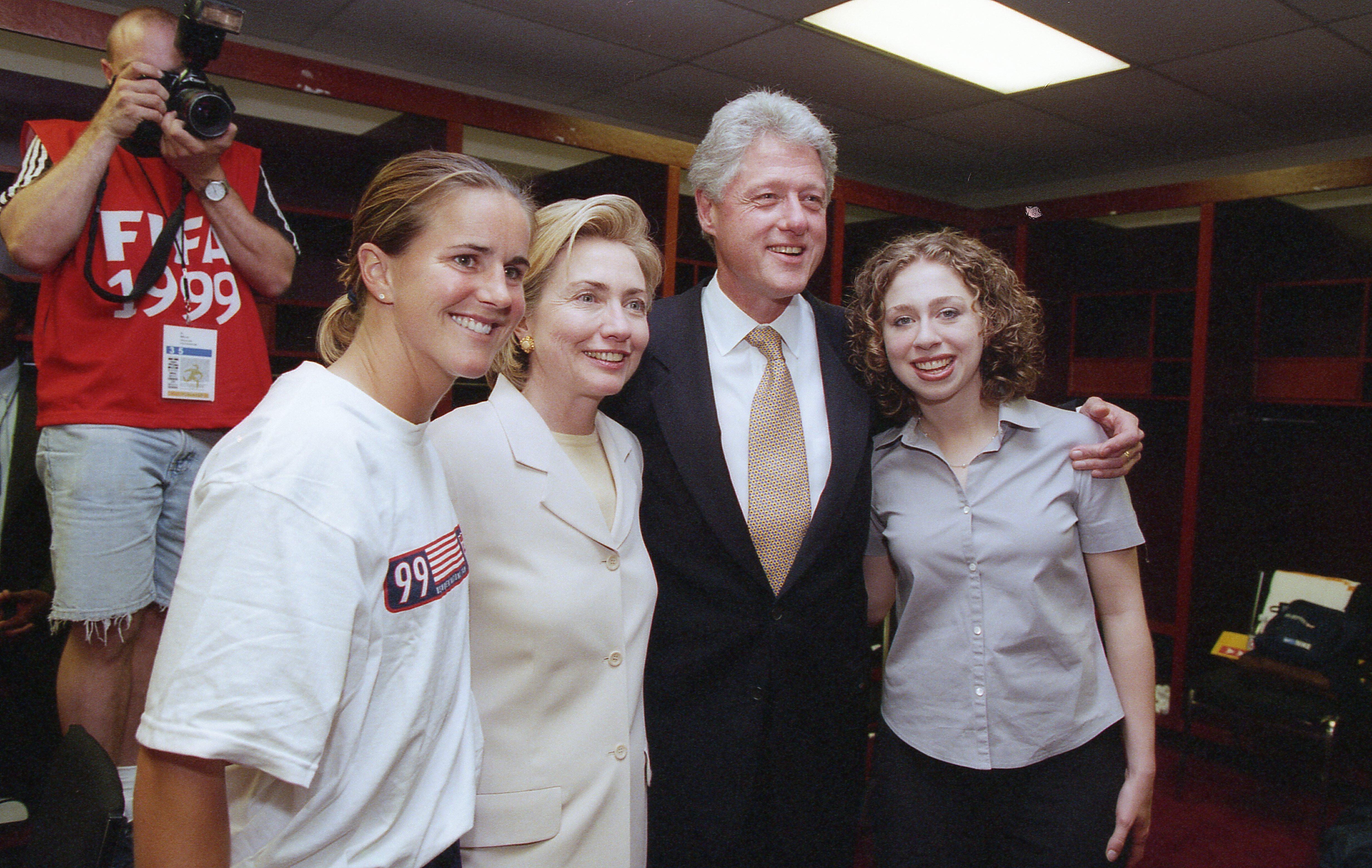 A photograph shows a Brandi Chastain wearing a white graphic t-shirt posing alongside Hillary Clinton, Bill Clinton, and Chelsea Clinton inside a locker room while a photographer stands in the background.