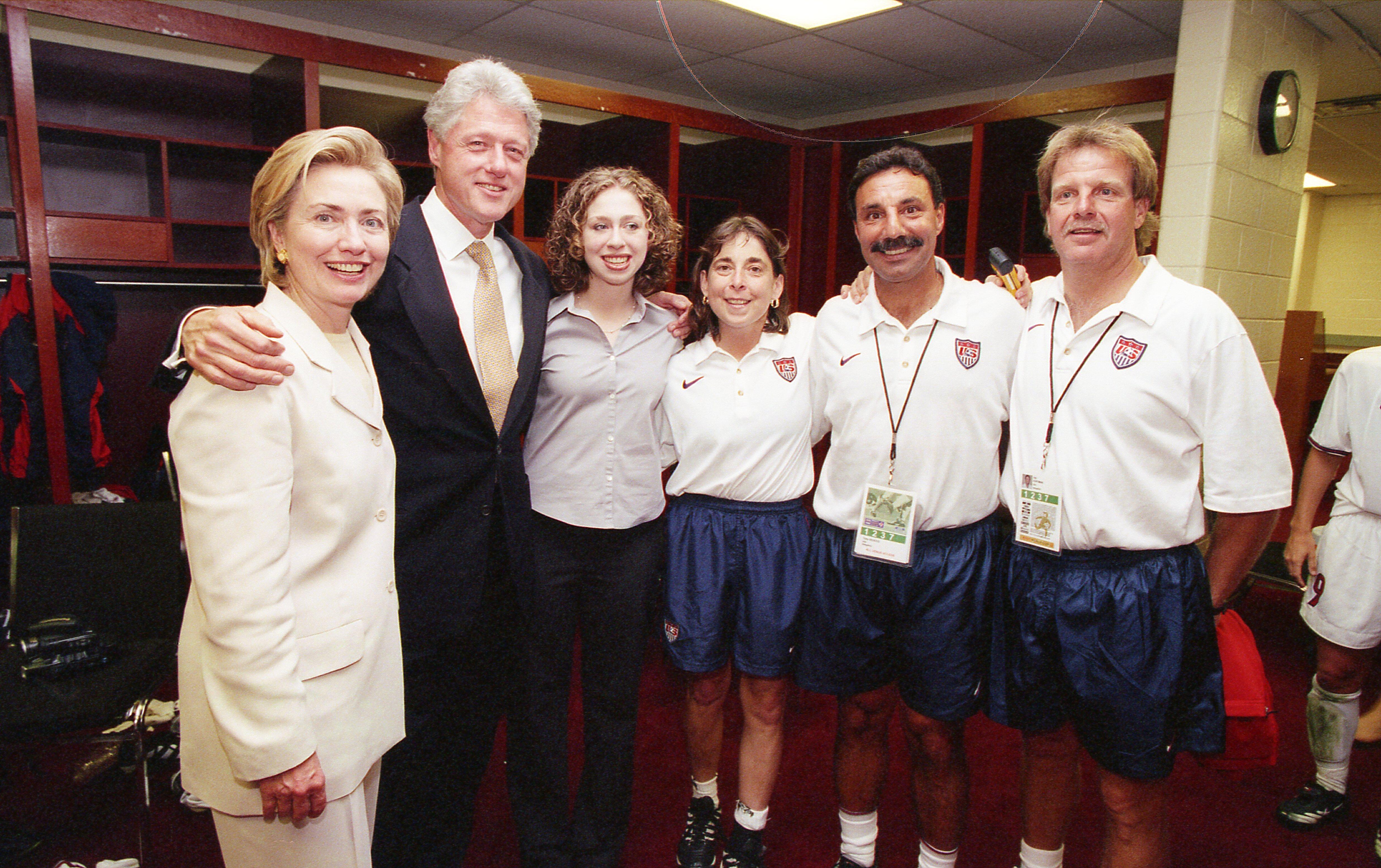 A photograph shows Hillary Clinton, Bill Clinton, and Chelsea Clinton smiling while posing in a locker room with three individuals wearing white United States Soccer Federation polo shirts and blue athletic shorts.