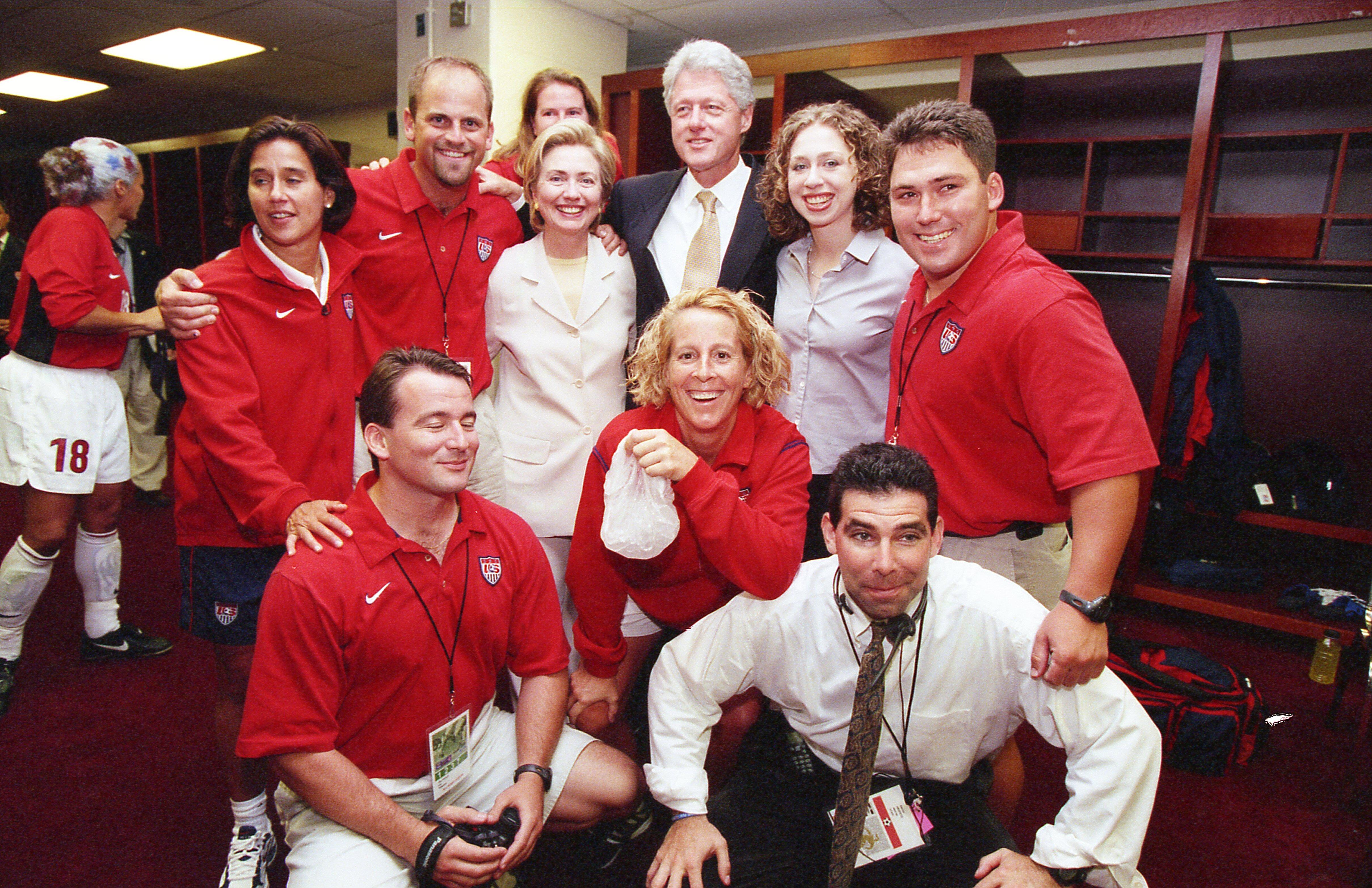 A photograph shows Hillary Clinton, Bill Clinton, and Chelsea Clinton posing for a group photo inside a locker room with several staff members wearing matching red United States Soccer Federation polo shirts and jackets.