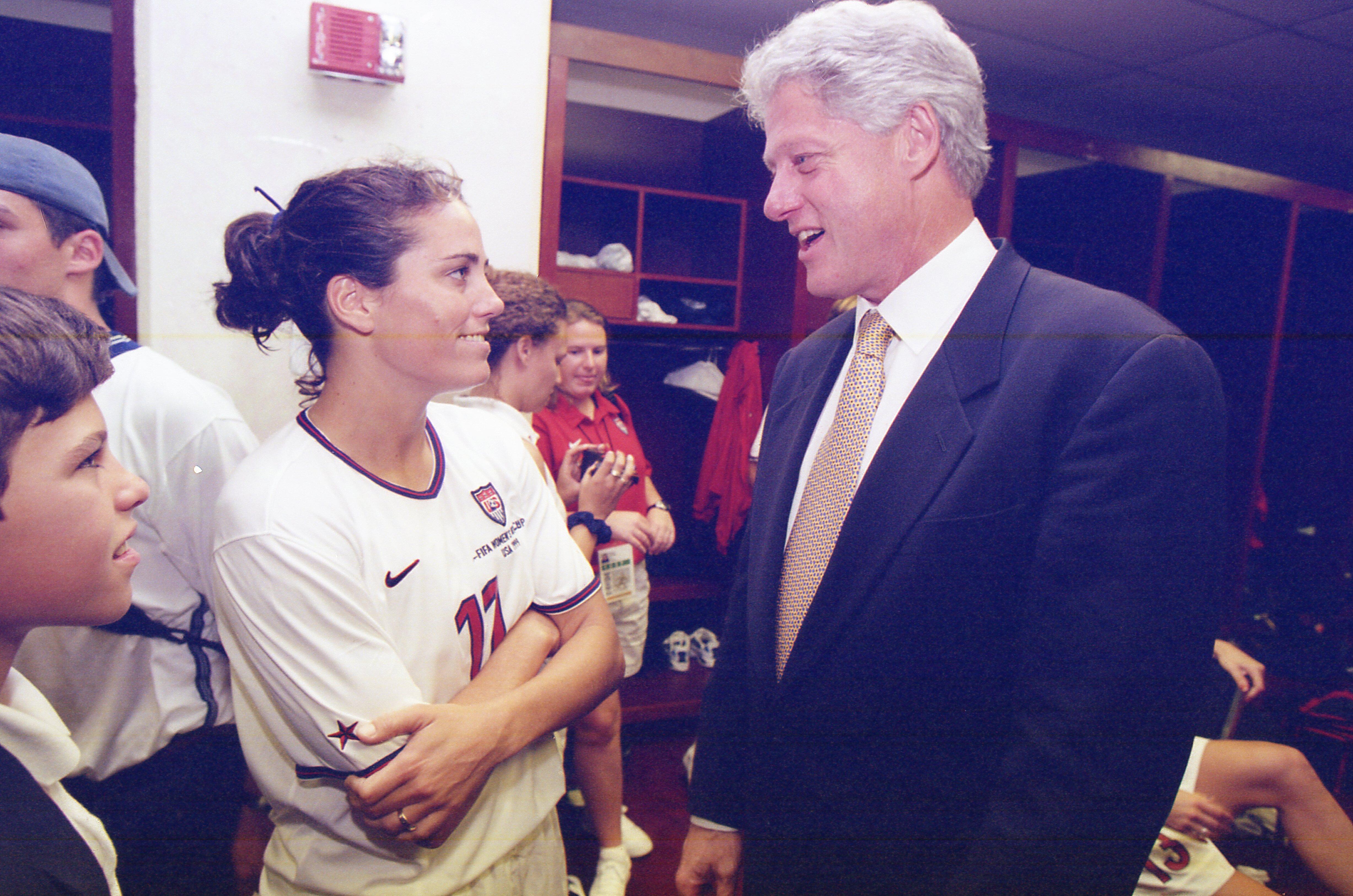 A photograph shows Bill Clinton smiling and speaking with U.S. soccer player Julie Foudy, who is wearing her white number 11 jersey, inside a locker room after a FIFA Women's World Cup match.