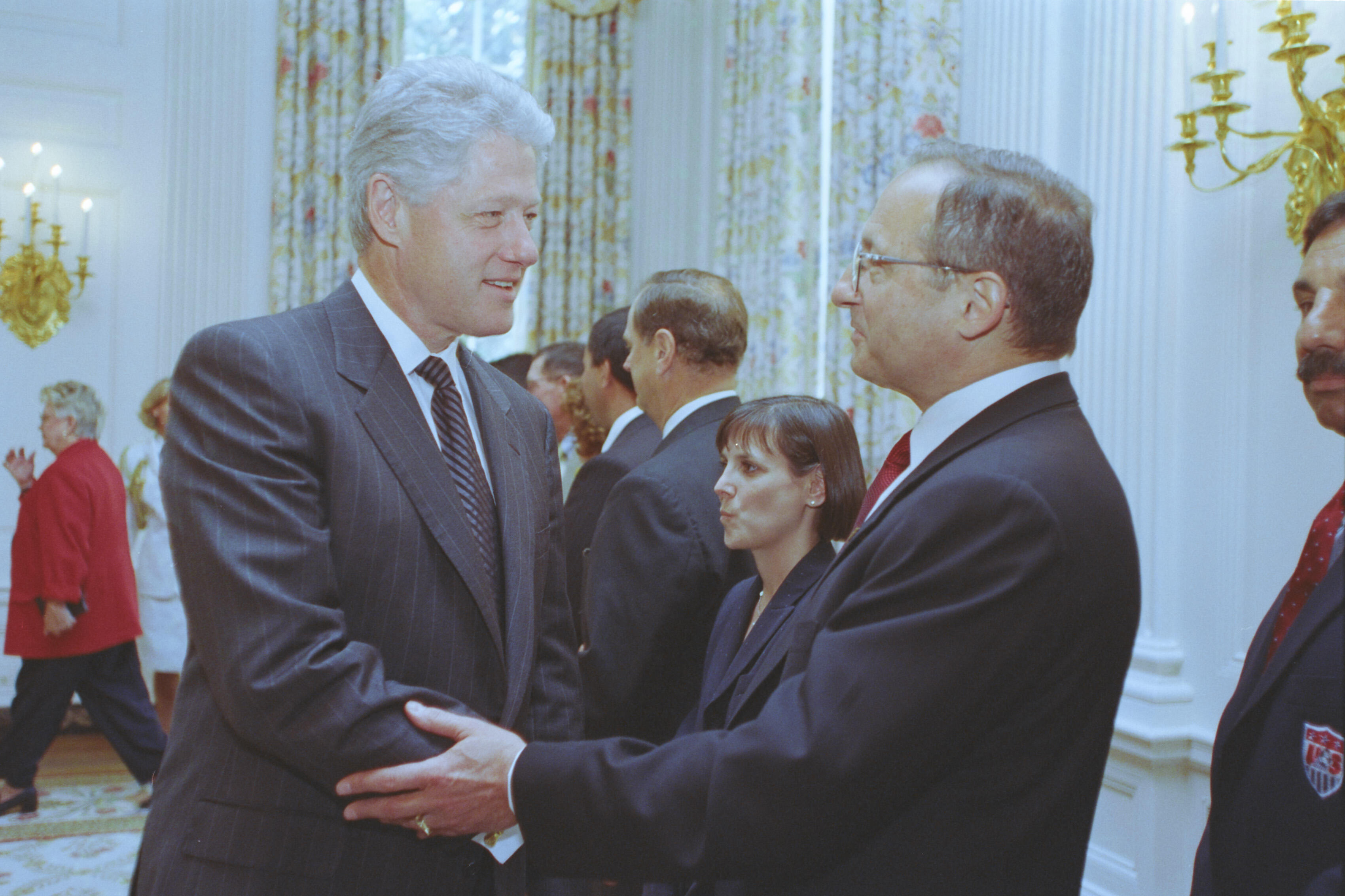 A medium shot captures former U.S. President Bill Clinton smiling and shaking hands with a man wearing glasses and a dark suit. A woman and several other men in business attire stand in the background of the elegant, well-lit room adorned with floral curtains and ornate wall sconces.