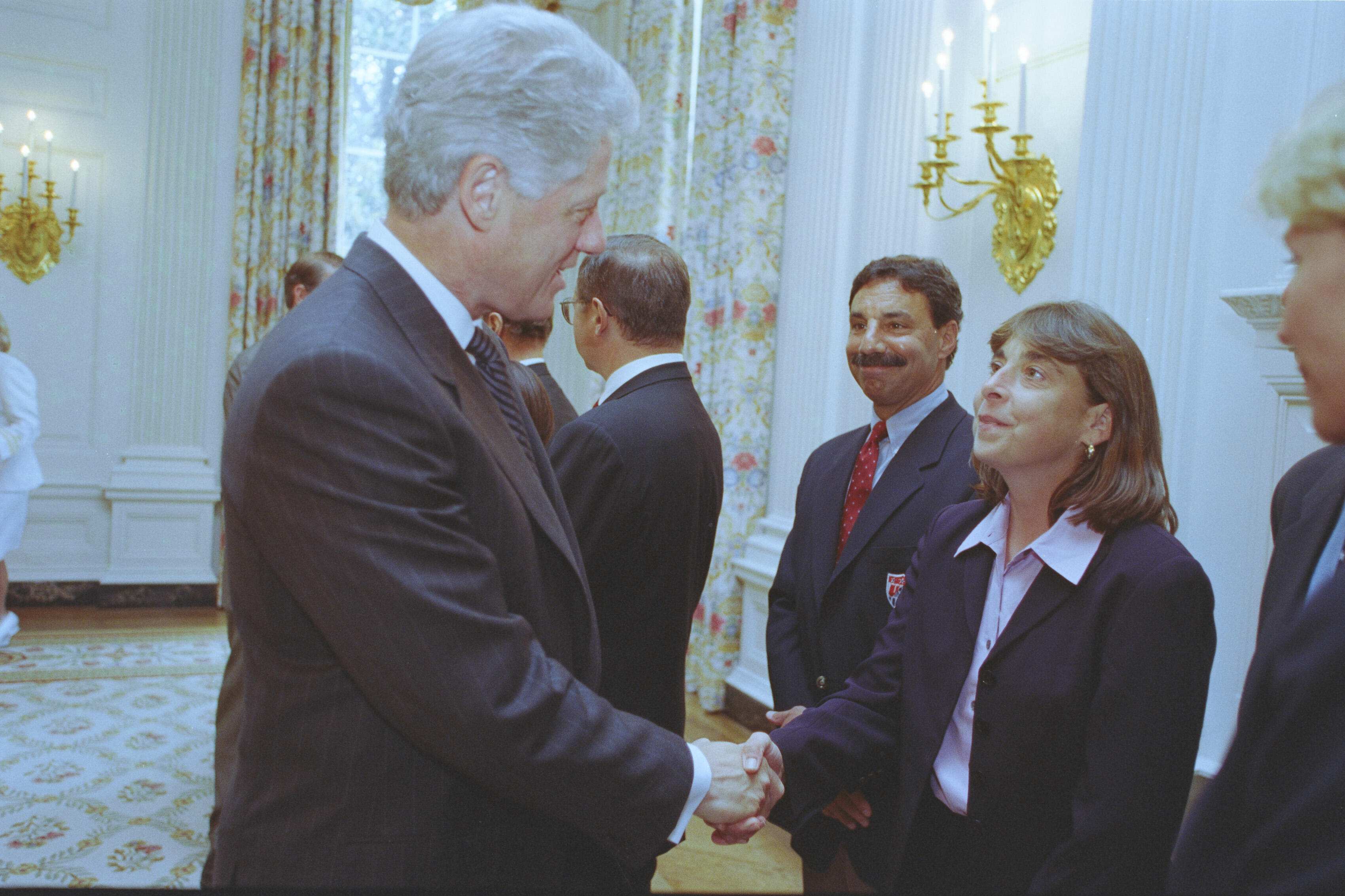 A medium shot captures former U.S. President Bill Clinton from the side profile as he smiles and shakes hands with a woman wearing a dark blazer over a light pink button-down shirt. A man with a mustache stands in a navy blazer behind her, while other guests and decorative gold sconces are visible in the background of the elegant room.