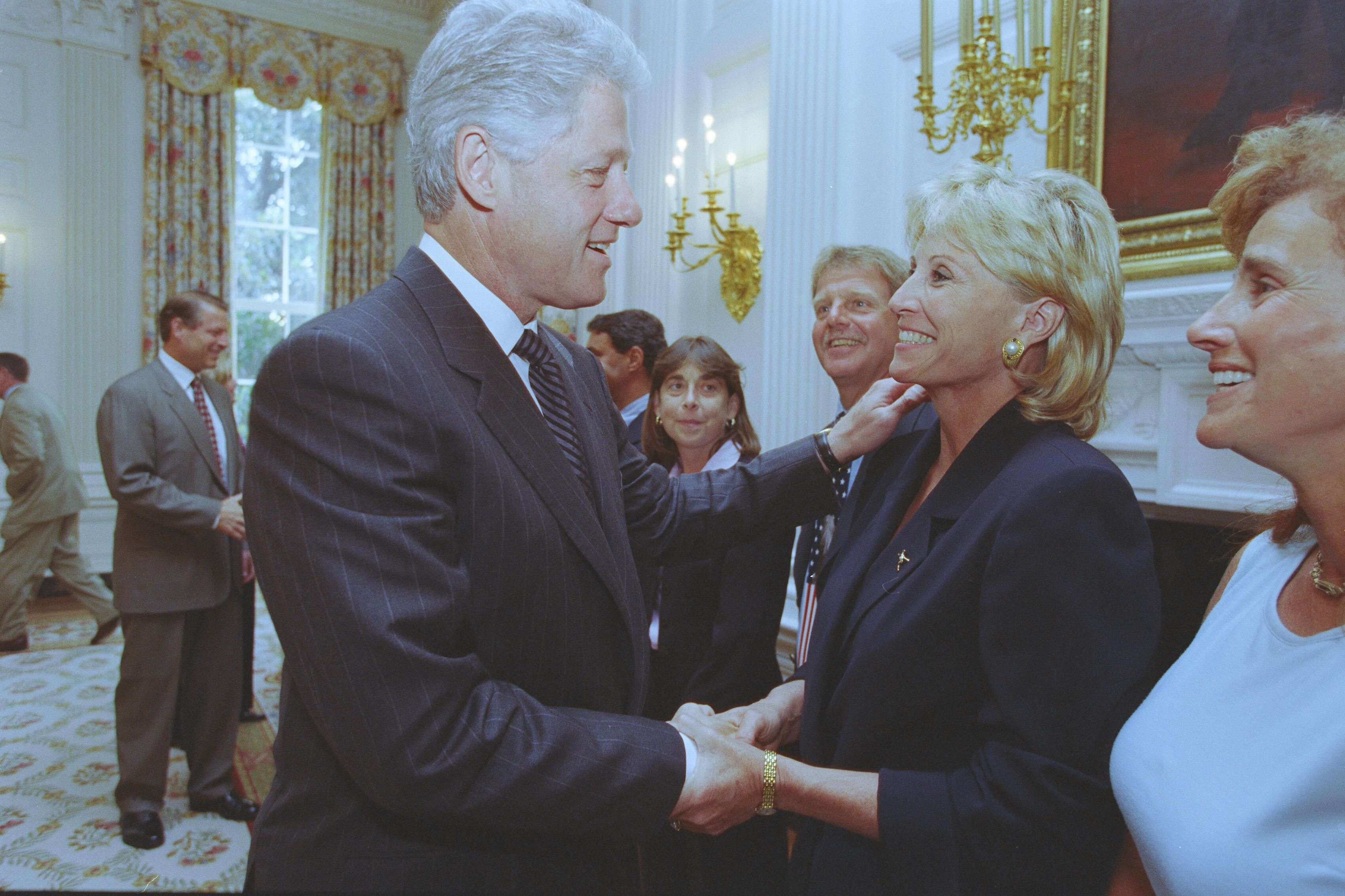 A medium shot captures former U.S. President Bill Clinton smiling as he shakes hands with a blonde woman wearing a dark blazer and a gold watch. With his other hand, he gently touches her shoulder as they interact. In the background, other guests look on, including Vice President Al Gore on the left, set against an elegant room with tall windows and decorative lighting fixtures.