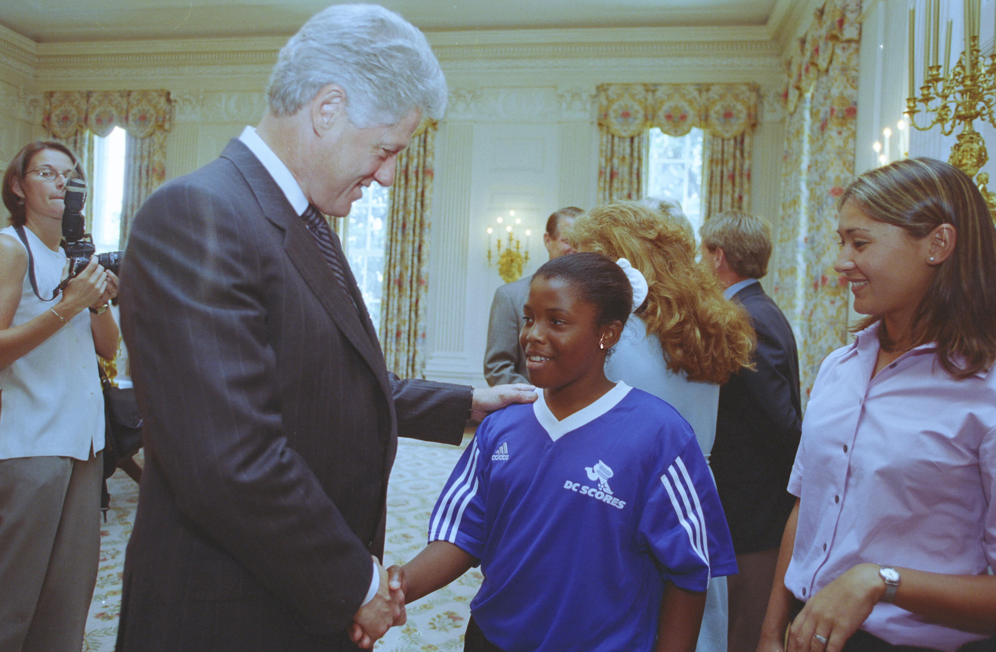 A medium shot shows former U.S. President Bill Clinton smiling as he shakes hands with a young Black girl wearing a blue Adidas sports jersey that reads "DC SCORES." He places his other hand gently on her shoulder. On the left, a photographer holds a camera, while another woman smiles on the right side of the frame, all set within a brightly lit, formal room with patterned curtains.