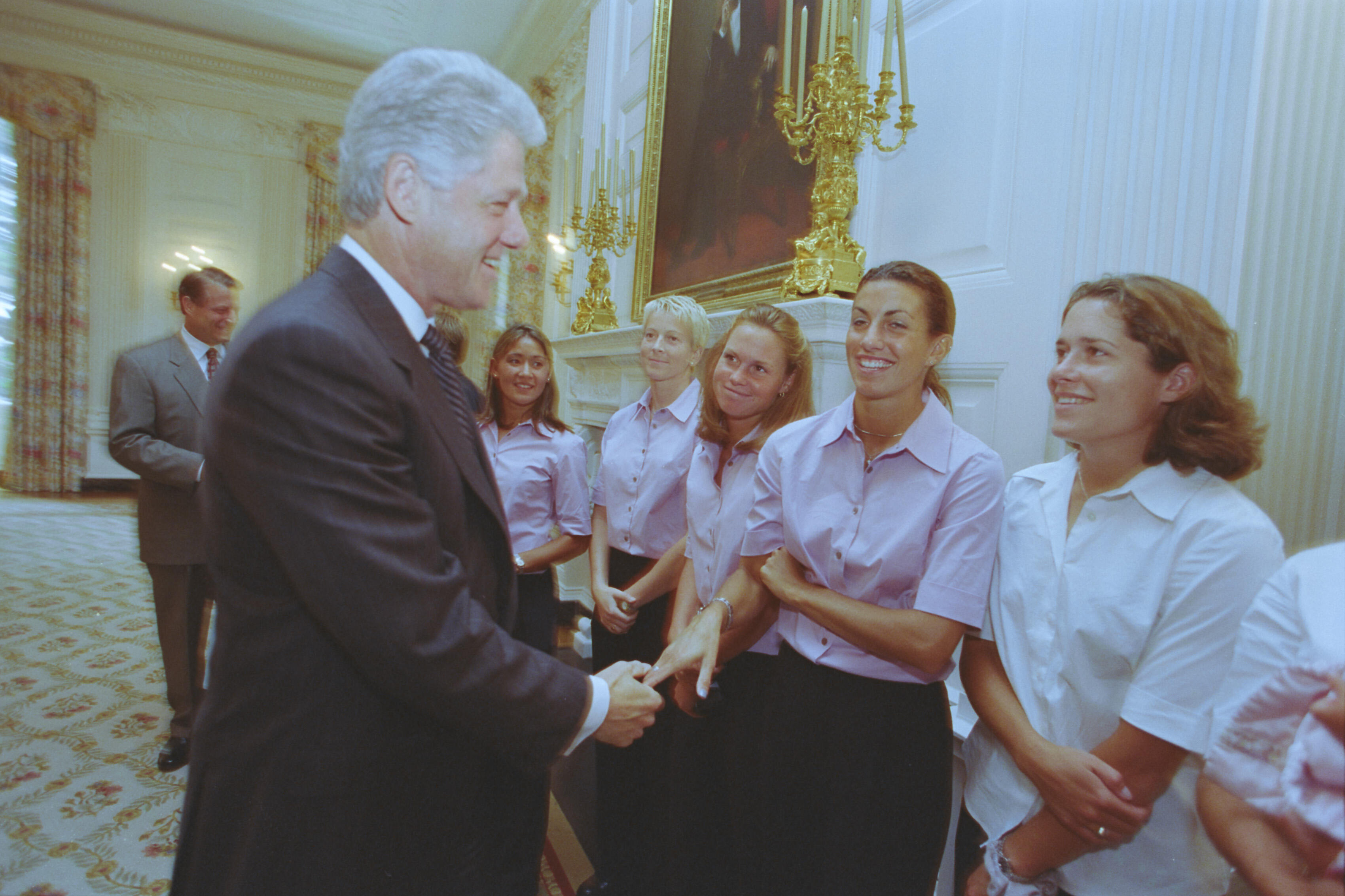 A medium shot captures former U.S. President Bill Clinton smiling and shaking hands with one of several women lined up in a well-lit room [1]. The women are smiling, with four wearing matching light pink short-sleeved button-down shirts and dark pants, while another wears a white button-down shirt. In the background, Vice President Al Gore stands on the left, and a large gold candelabra hangs on the ornate white wall.