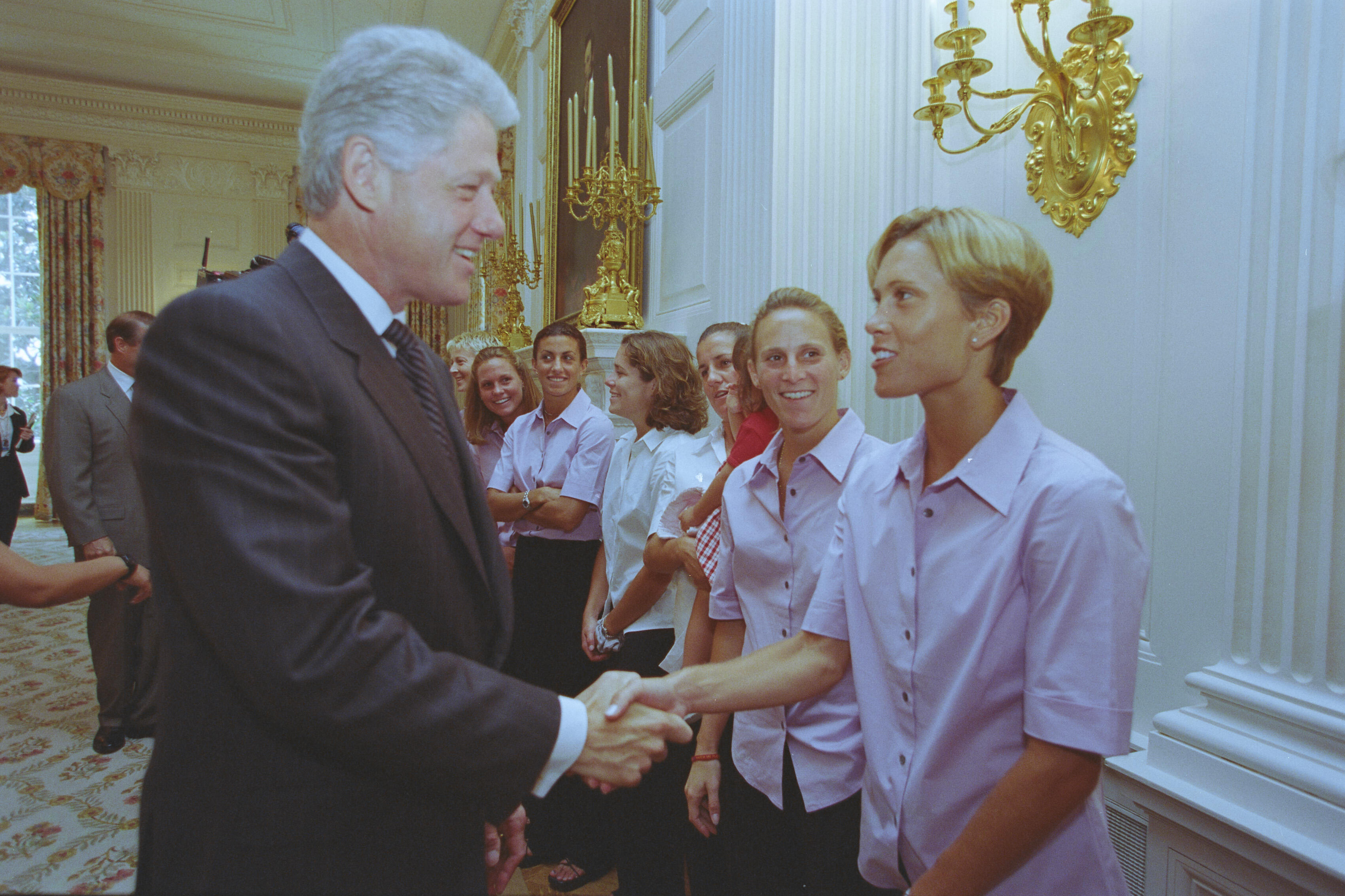 A medium shot captures former U.S. President Bill Clinton smiling and shaking hands with a woman with short blonde hair who is part of a line of women. Most of the women are smiling and wearing matching light pink short-sleeved button-down shirts with dark pants. On the far left, Vice President Al Gore is visible in a grey suit, while an ornate gold candelabra sconce decorates the white paneled wall on the right.