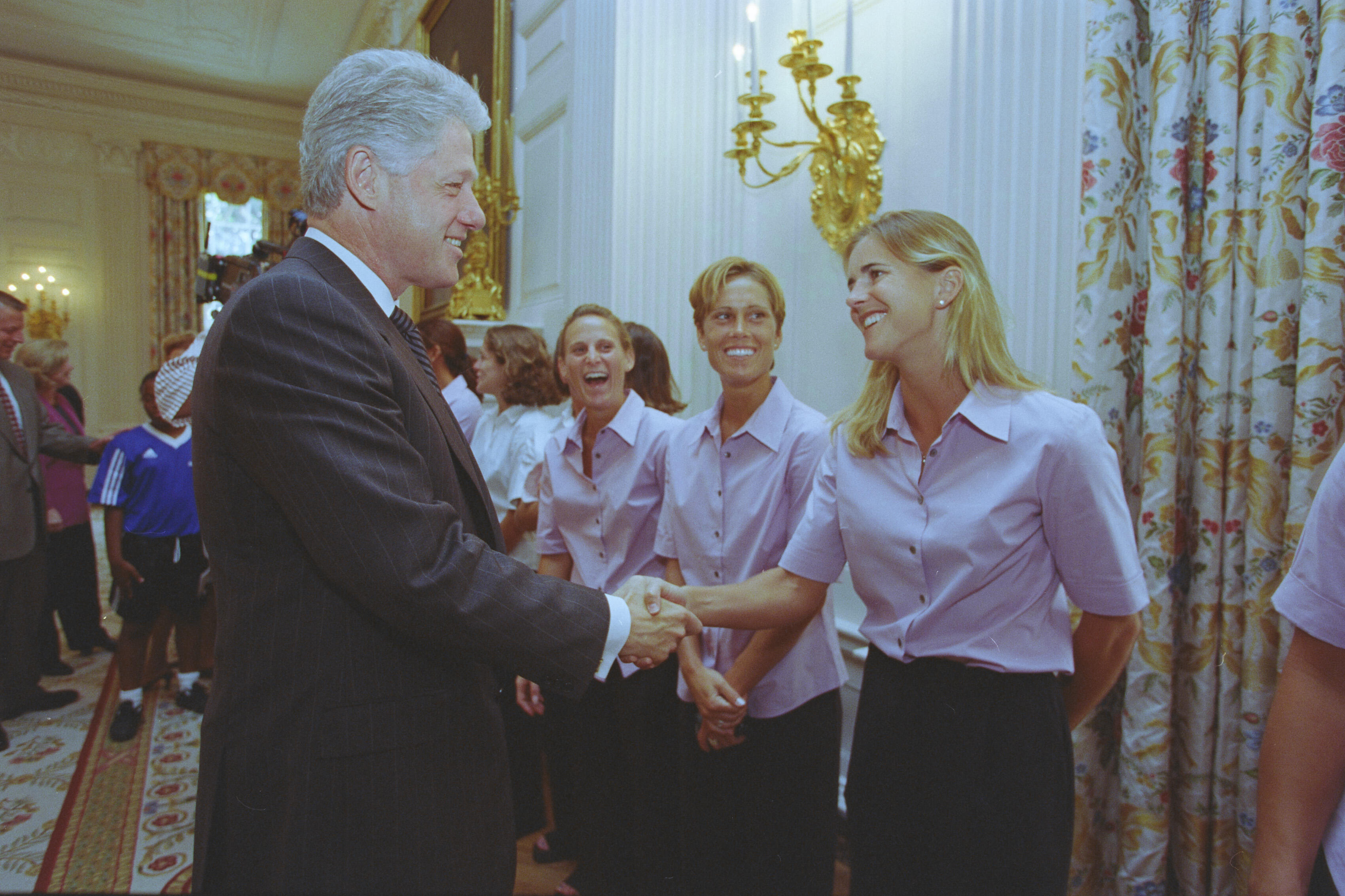 A medium shot captures former U.S. President Bill Clinton smiling as he shakes hands with a smiling, blonde Brandi Chastain. She stands in a line with other women wearing matching light pink short-sleeved button-down shirts and dark pants. In the background on the left, a young girl wears a blue sports jersey, while a gold wall sconce and patterned floral curtains decorate the elegant room.