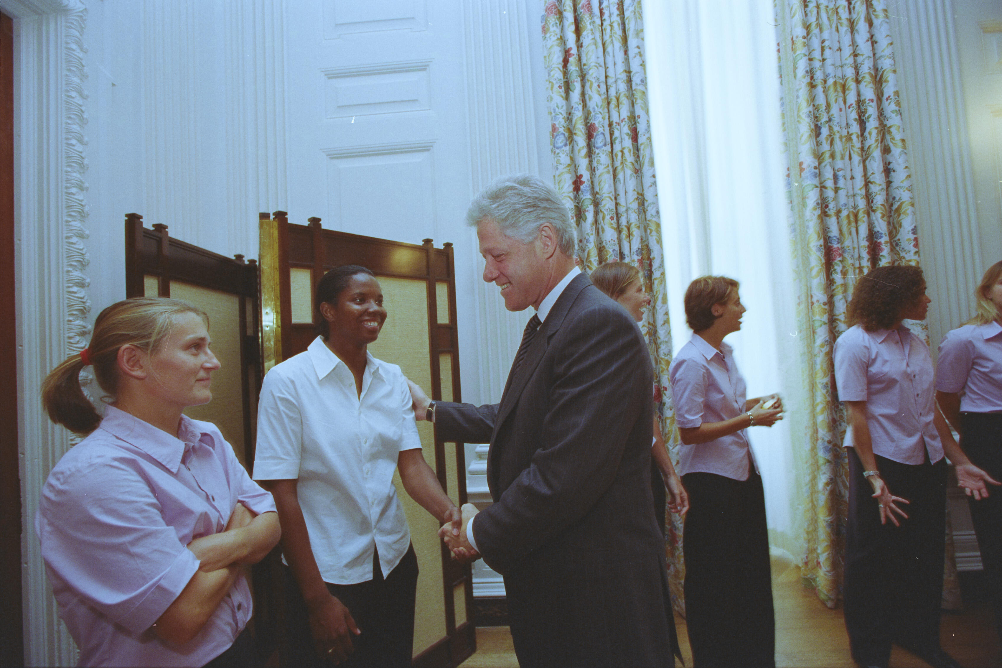 A medium shot captures former U.S. President Bill Clinton smiling as he shakes hands with a smiling Black woman wearing a white short-sleeved button-down shirt and dark pants. He places his other hand gently on her shoulder. On the left, another woman with her blonde hair in a ponytail stands with arms crossed, wearing a light pink shirt. In the background, several other women in matching light pink shirts look on near tall windows with floral-patterned curtains.