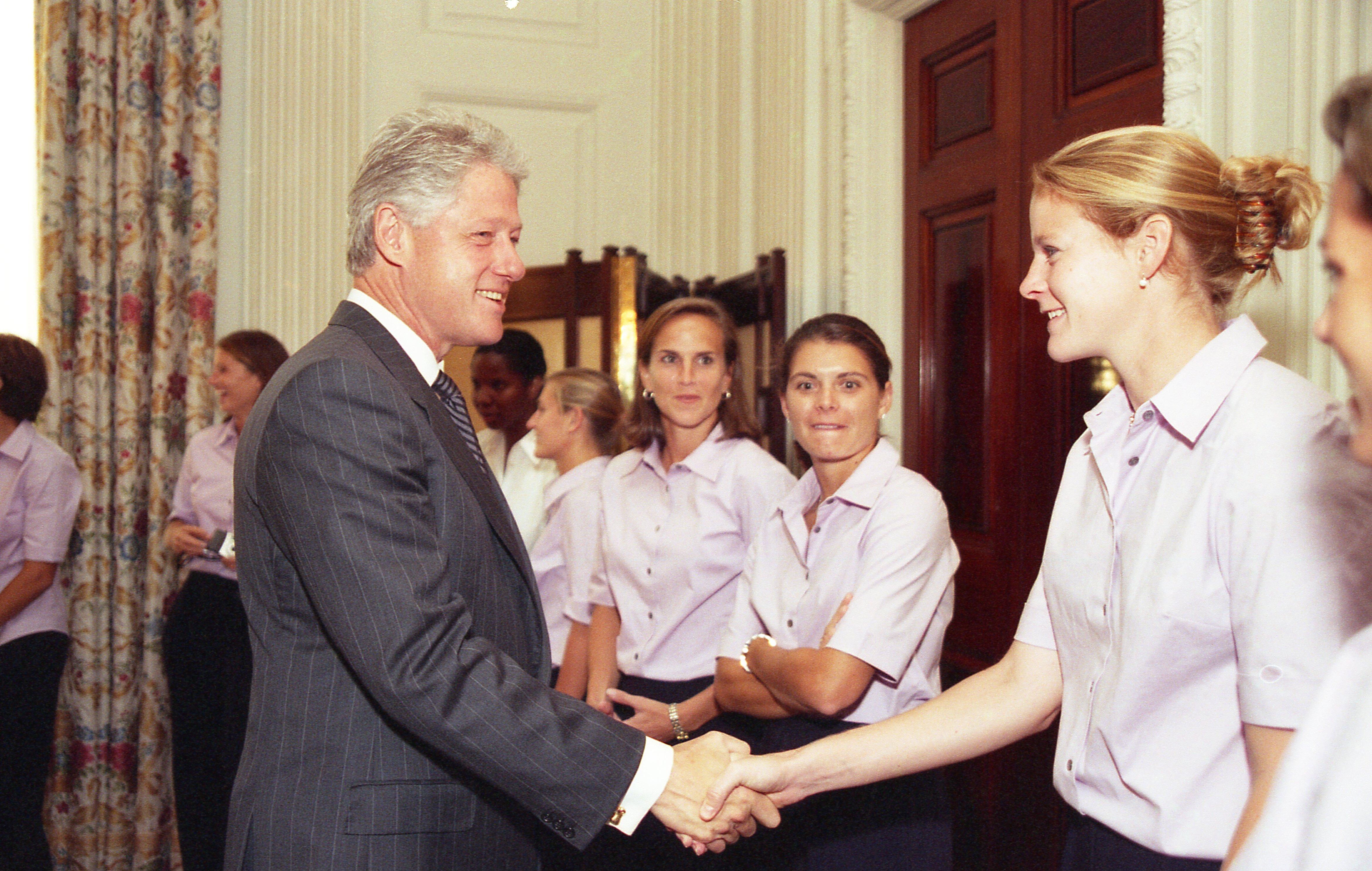 A medium shot captures former U.S. President Bill Clinton smiling as he shakes hands with a smiling blonde woman with her hair secured in a bun. She stands at the front of a line of women who are wearing matching light pink short-sleeved button-down shirts and dark pants. The greeting occurs near a large dark wood door frame, with floral-patterned curtains visible in the background on the far left.