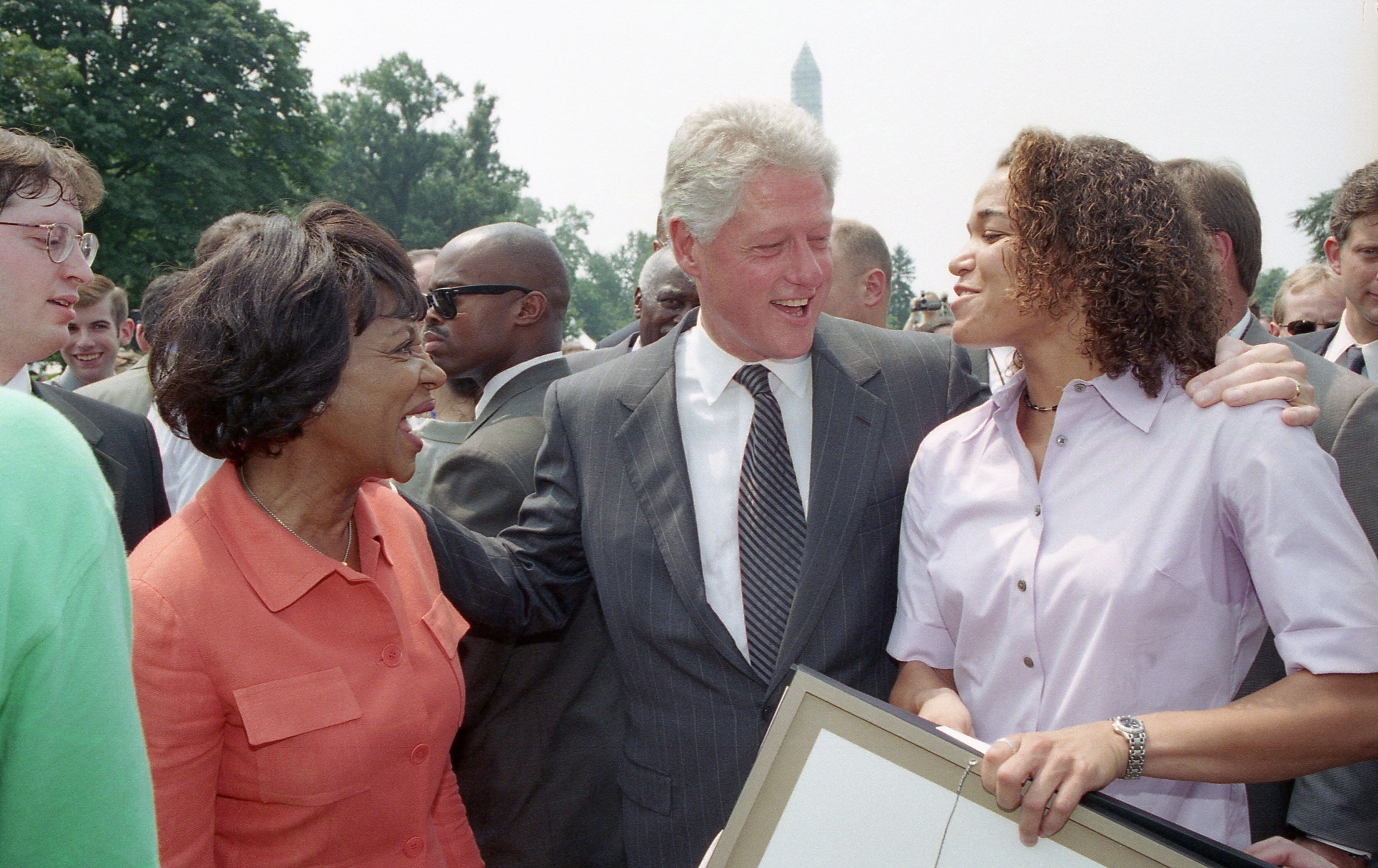 A medium shot captures former U.S. President Bill Clinton smiling and conversing outdoors with a woman in an orange button-down jacket on his right and a woman with curly hair in a light pink shirt on his left, placing his arm around her shoulder. The woman in the pink shirt holds a framed document or award, while a crowd of other people stands closely in the background under a cloudy sky, with the Washington Monument faintly visible in the far distance.