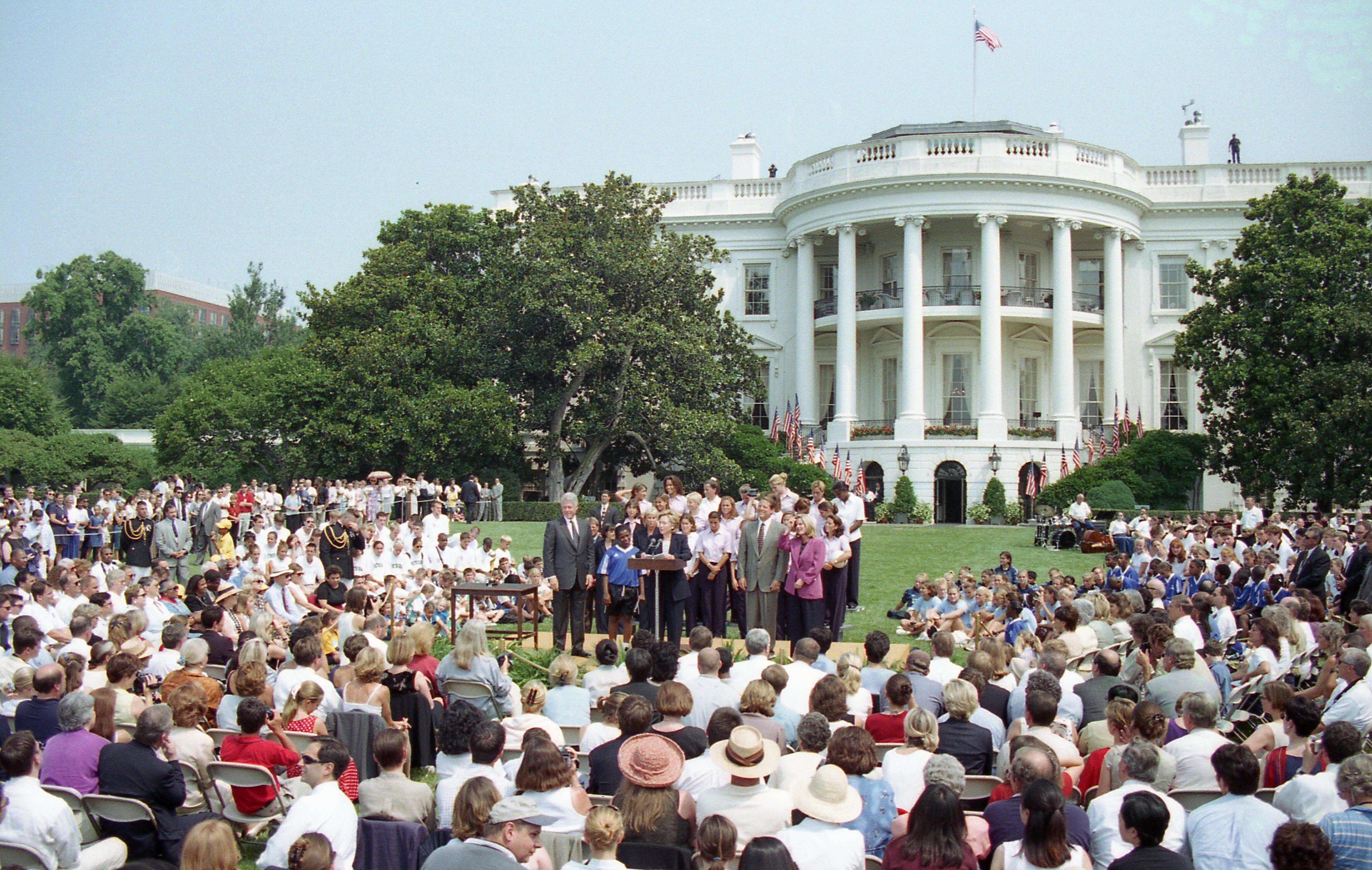 A large crowd of people sits on folding chairs and on the lawn of the White House South Portico, facing a small raised stage. On the stage, President Bill Clinton, First Lady Hillary Clinton, and several other officials and athletes stand at a podium. The iconic white, columned facade of the White House rises in the background under a clear sky, decorated with numerous American flags along its balcony railings. Large green trees flank the lawn, framing the gathering.