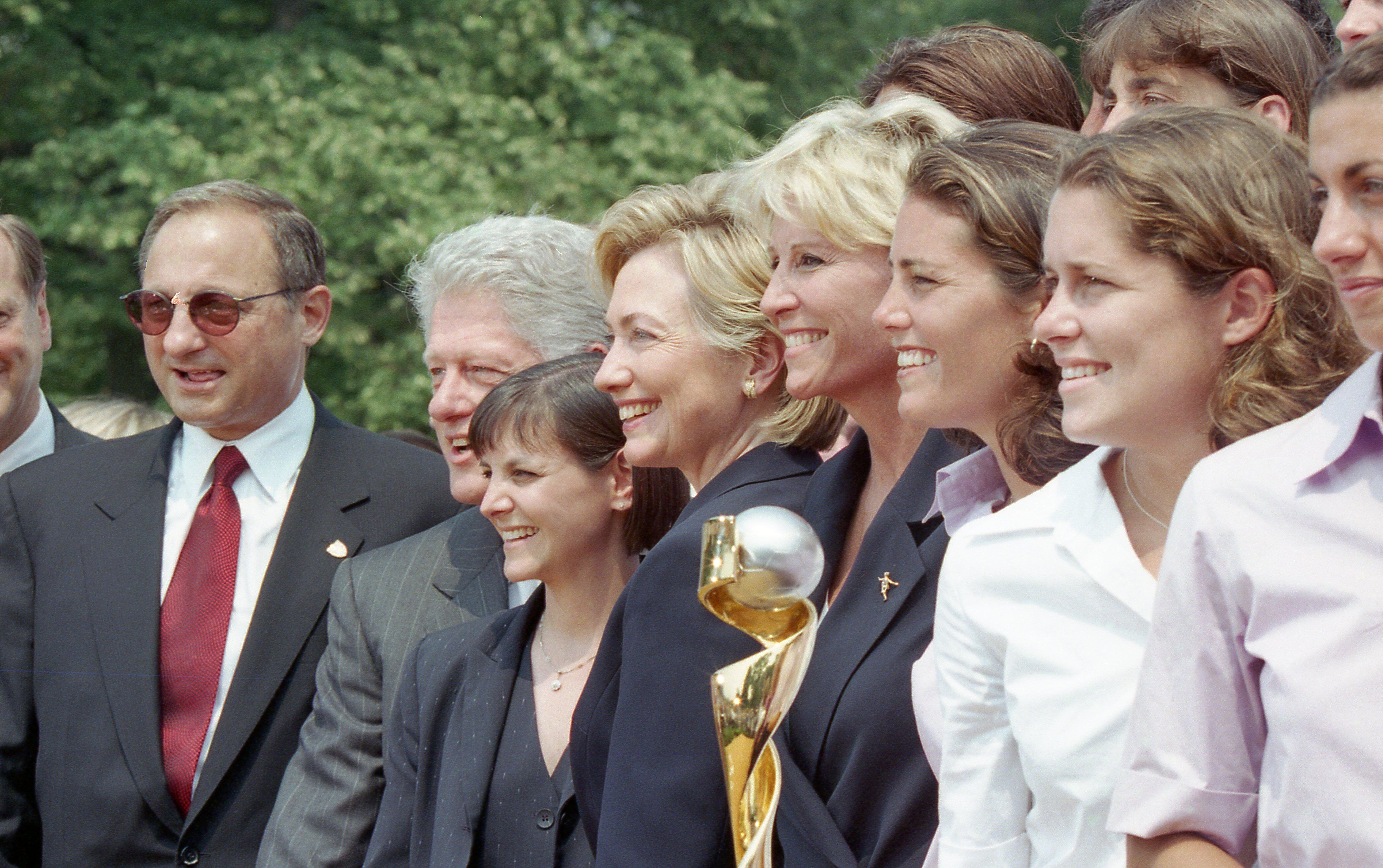A close-up photograph captures former President Bill Clinton and First Lady Hillary Clinton standing outside with a group of people, including members of the U.S. Women’s National Soccer Team.Hillary Clinton stands near the center, smiling in profile. To her left is former President Bill Clinton, also smiling. A gold and silver World Cup trophy stands prominently in the foreground. Several smiling women and a man in a suit and sunglasses stand alongside them against a background of green trees.