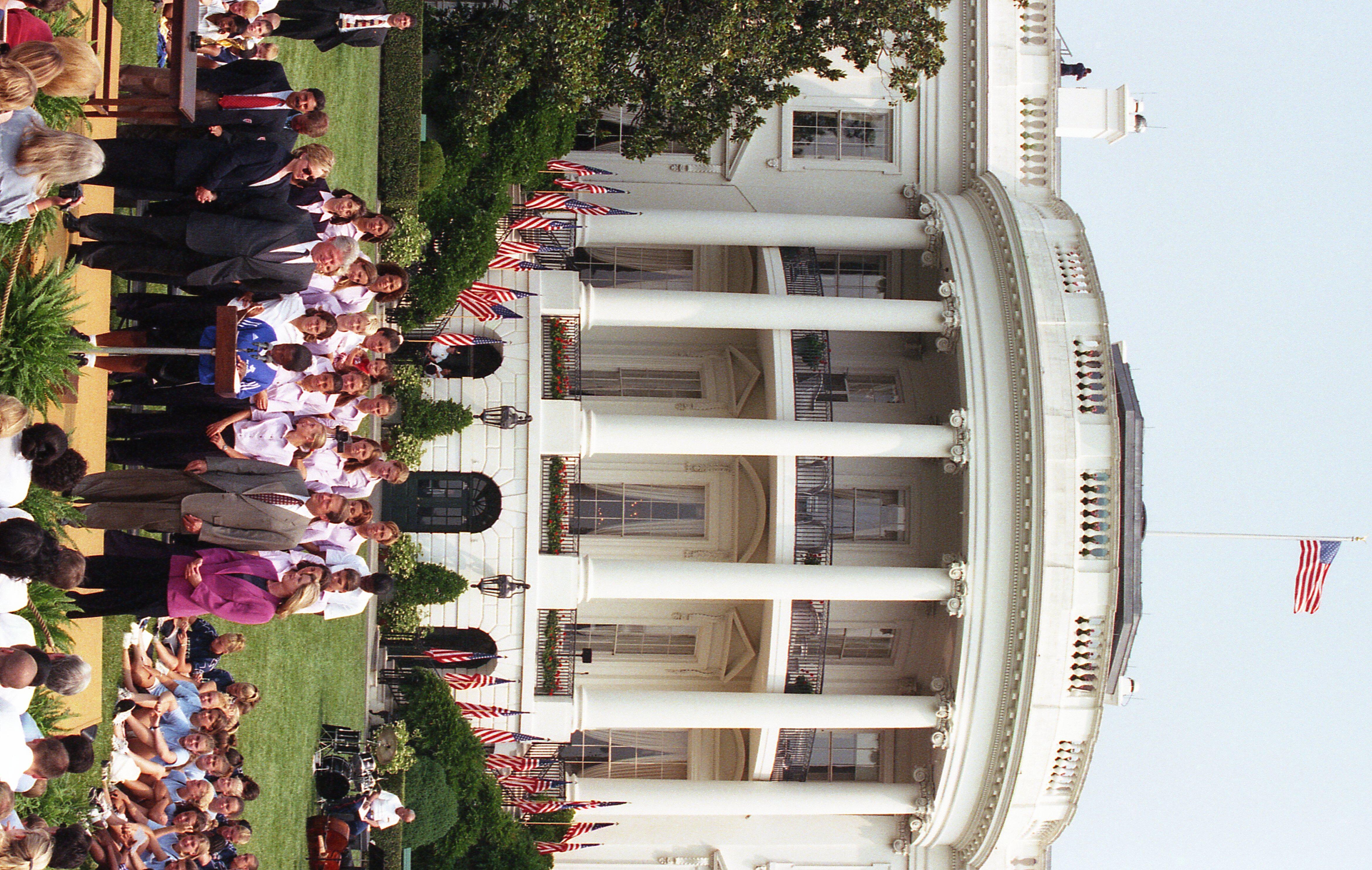 A wide shot captures the South Portico of the White House, where former U.S. President Bill Clinton and First Lady Hillary Clinton stand alongside Vice President Al Gore and Second Lady Tipper Gore on a small stage. They are gathered with a large group of women wearing matching light pink shirts and dark pants, and a young girl in a blue "DC SCORES" sports jersey speaks into microphones at a wooden podium. A crowd sits on the South Lawn grass in front of the stage, flanked by multiple American flags.