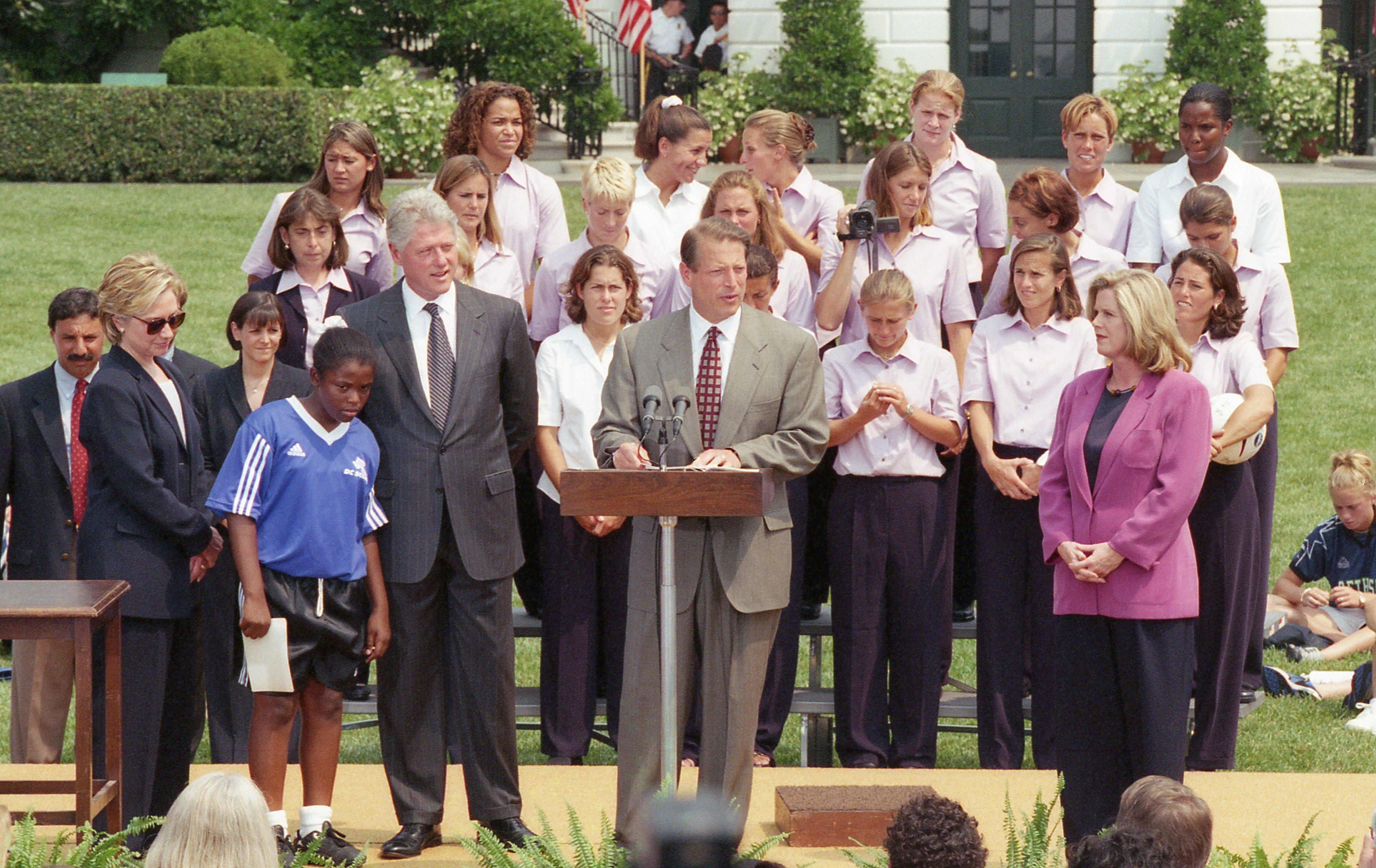 A wide shot captures Vice President Al Gore speaking at a podium on an outdoor stage at the White House, surrounded by political figures and the U.S. Women’s National Soccer Team.To the left of the podium stands President Bill Clinton next to a young girl in a blue and black soccer uniform. Further left, First Lady Hillary Clinton looks downward. Tipper Gore stands to the right of the podium wearing a purple blazer. Behind them, members of the soccer team stand in rows wearing matching light pink shirts, al
