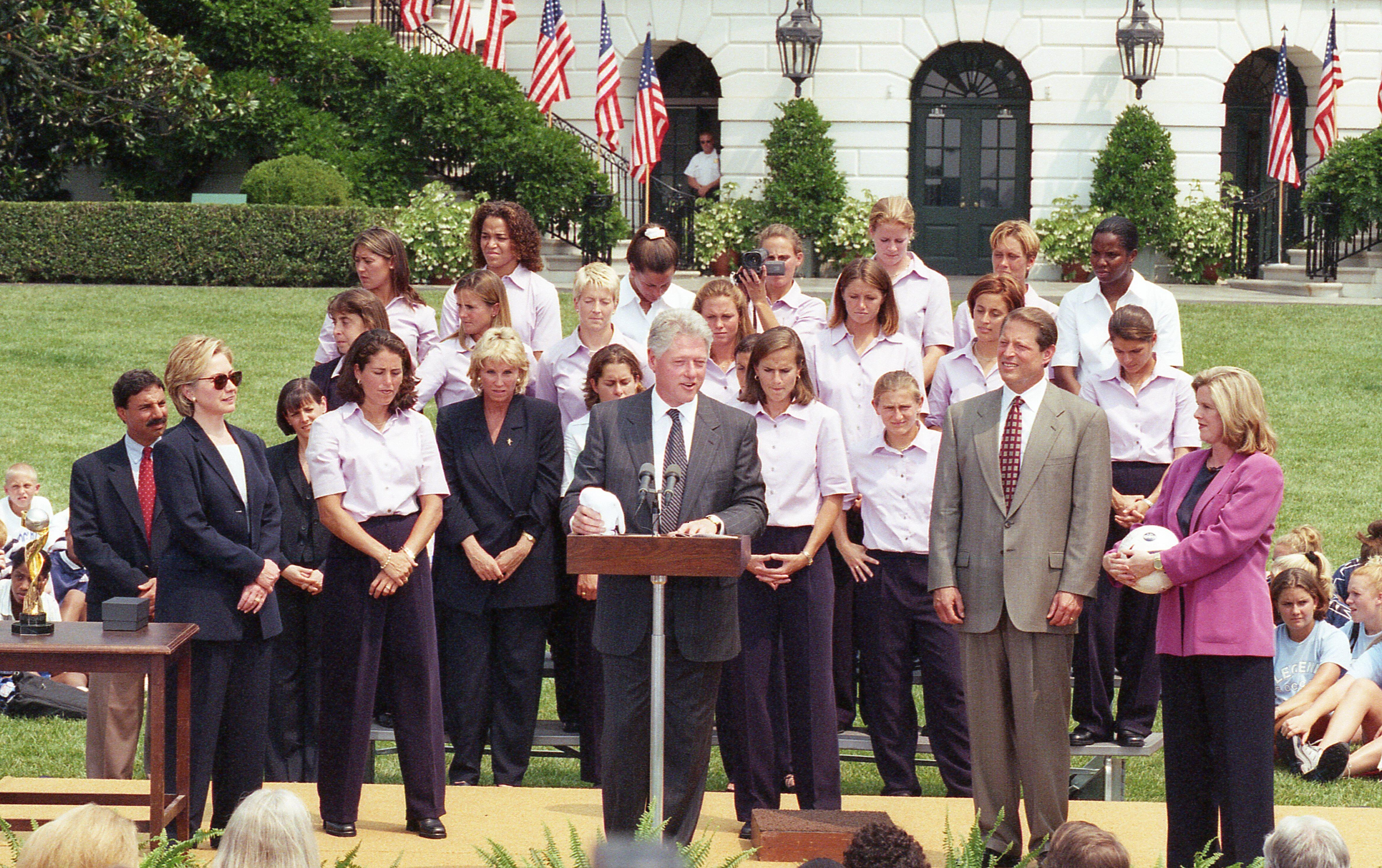 An official outdoor ceremony at the White House features Bill Clinton speaking at a wooden podium in the center. He wears a dark grey suit and a striped tie. To his right, Al Gore stands in a light grey suit and a patterned red tie, next to Tipper Gore who is holding a white soccer ball and wearing a purple blazer. To the left of the podium stands Hillary Clinton wearing sunglasses and a dark pantsuit. A large group of young women, wearing matching light purple button-down shirts and dark pants,.