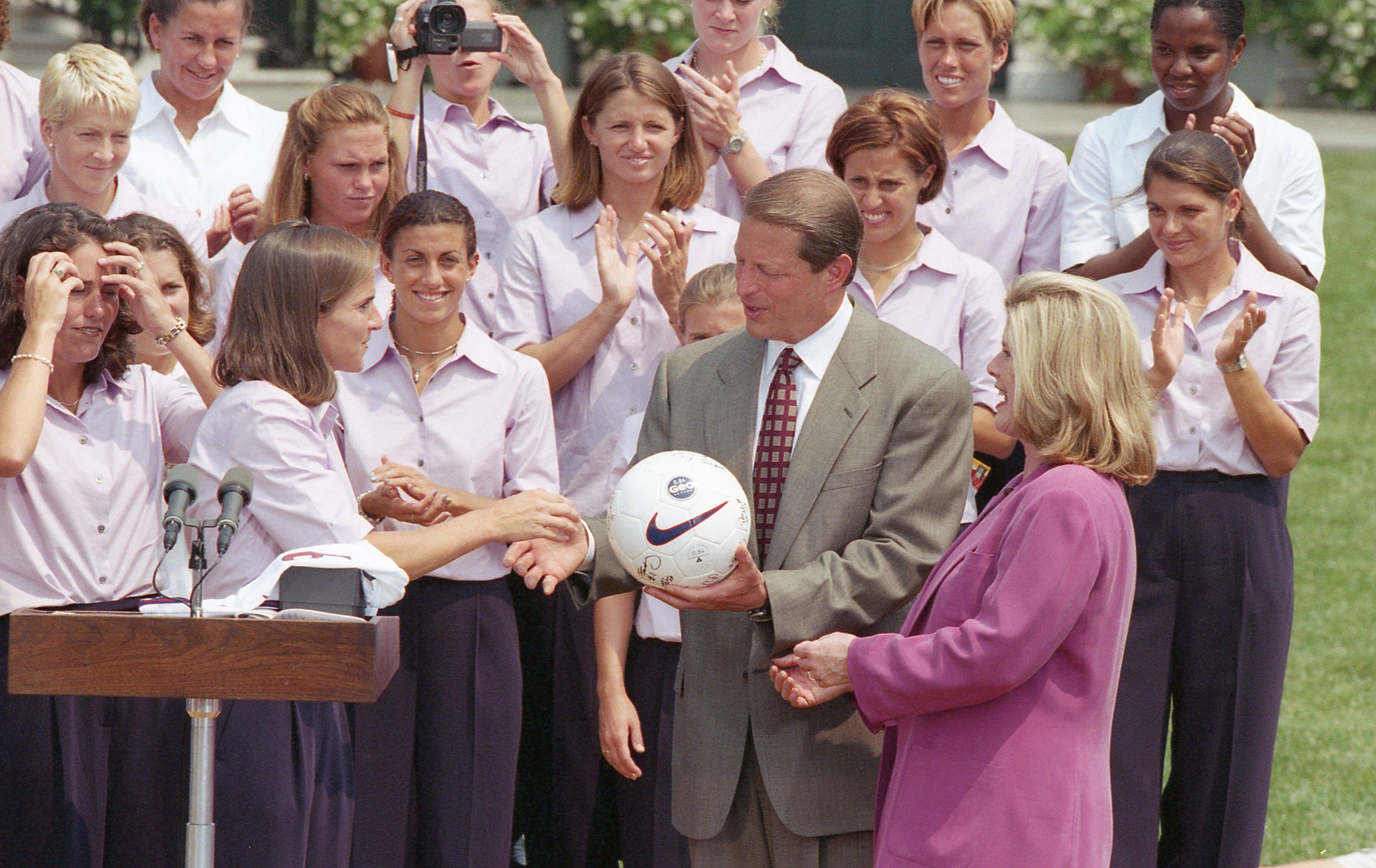 An outdoor ceremony at the White House shows Al Gore holding a white Nike soccer ball in the center. He wears a grey suit jacket and a red patterned tie. To his right, Tipper Gore stands in profile, wearing a magenta blazer and smiling toward the team. On the left side of the frame, several members of a women's soccer team shake hands near a wooden podium with two microphones. The team members wear matching light purple button-down shirts and dark pants, and many are smiling and clapping in the background.