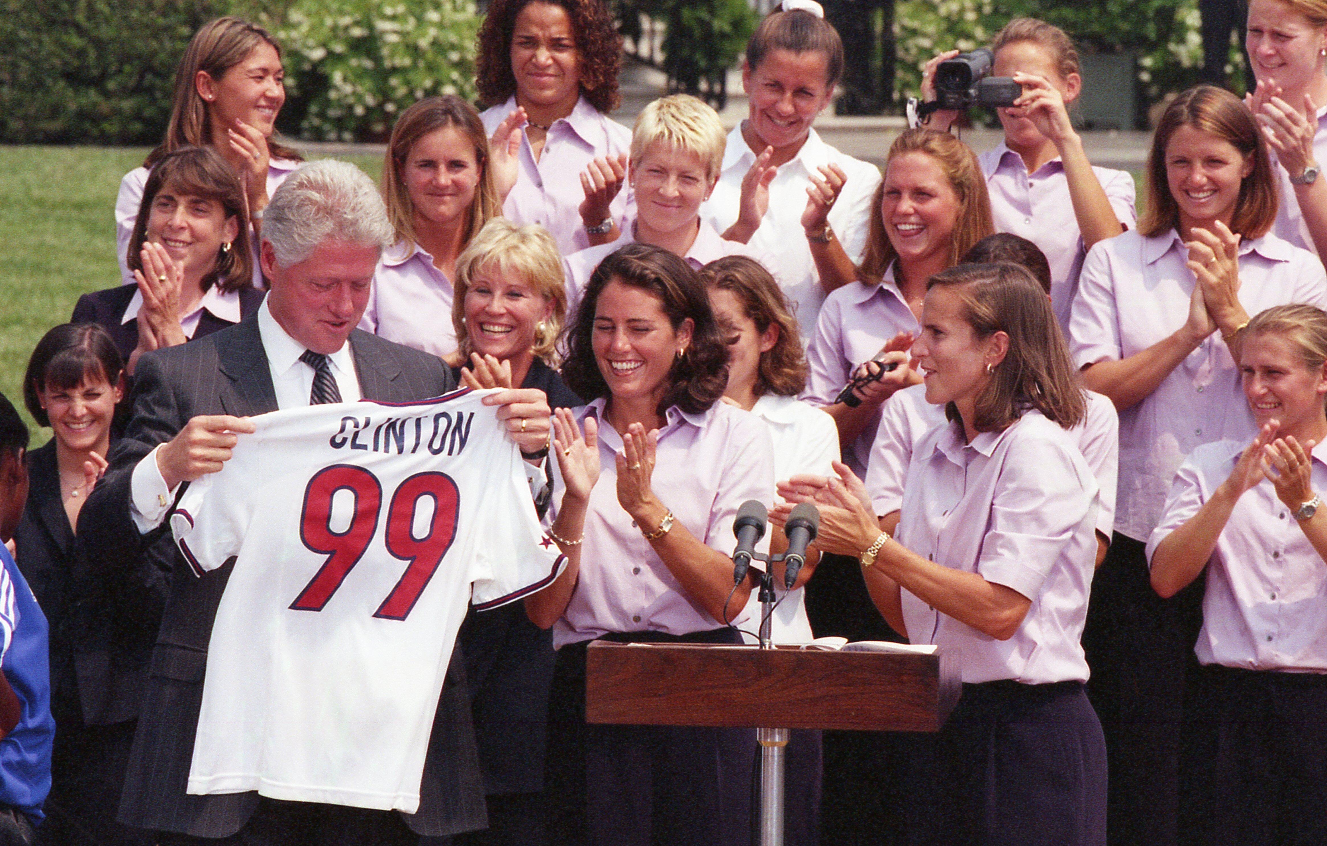 An outdoor White House ceremony features Bill Clinton holding up a white soccer jersey with the name "CLINTON" and the number "99" printed on the back. He stands on the left in a dark pinstripe suit. Two female soccer players stand next to him behind a wooden podium, smiling and clapping. A large group of team members, dressed in matching light purple button-down shirts, are gathered closely behind them, clapping and cheering in bright daylight.