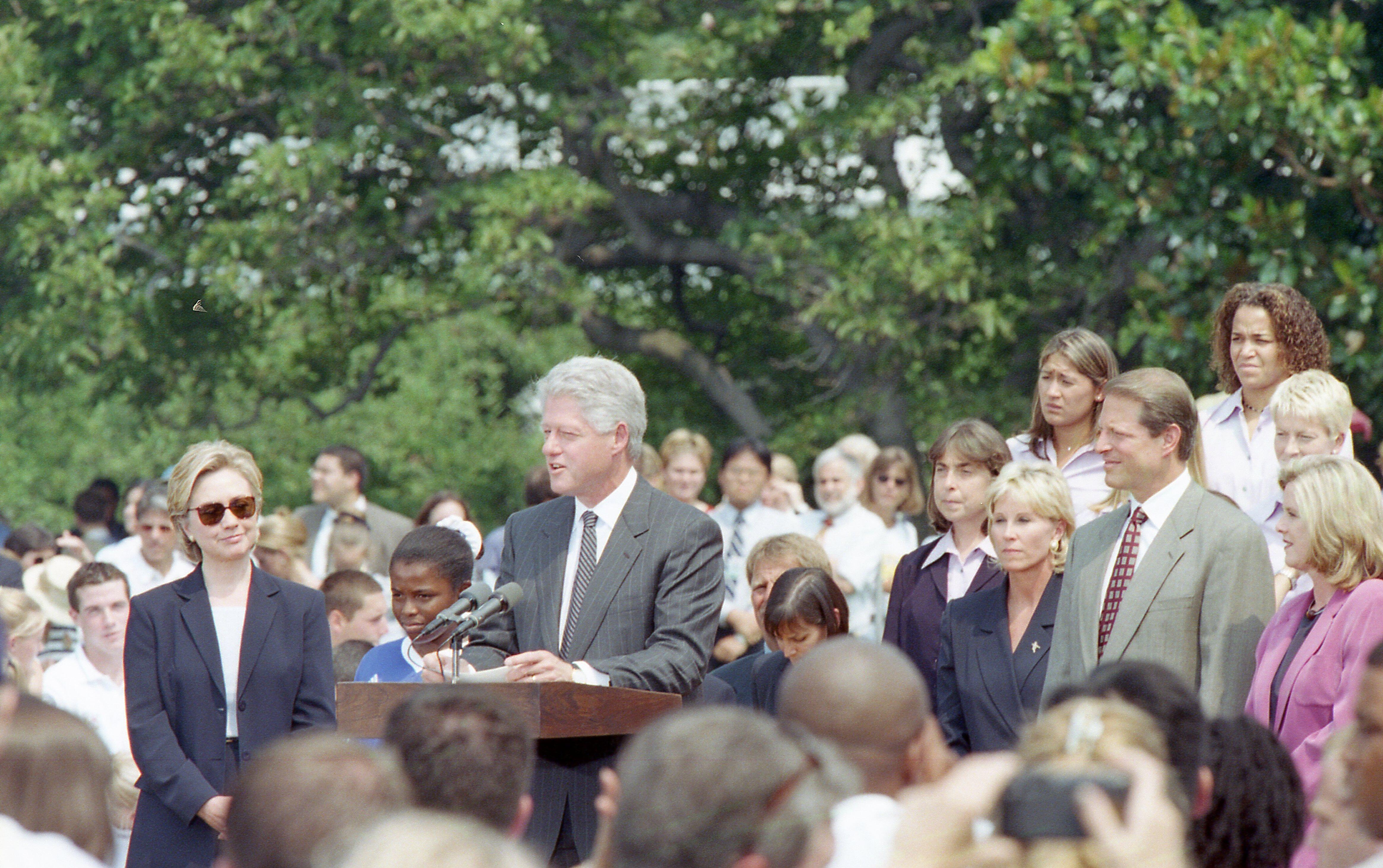 An outdoor White House ceremony features Bill Clinton speaking at a wooden podium in the center, wearing a dark pinstripe suit and striped tie. To his left stands Hillary Clinton in a dark pantsuit and sunglasses, next to a young boy in a blue uniform. To his right, Al Gore stands in a light grey suit and patterned red tie, flanked by Tipper Gore in a purple blazer and another blonde woman in a dark jacket. A crowd of onlookers and several team members in light purple shirts stand behind them.