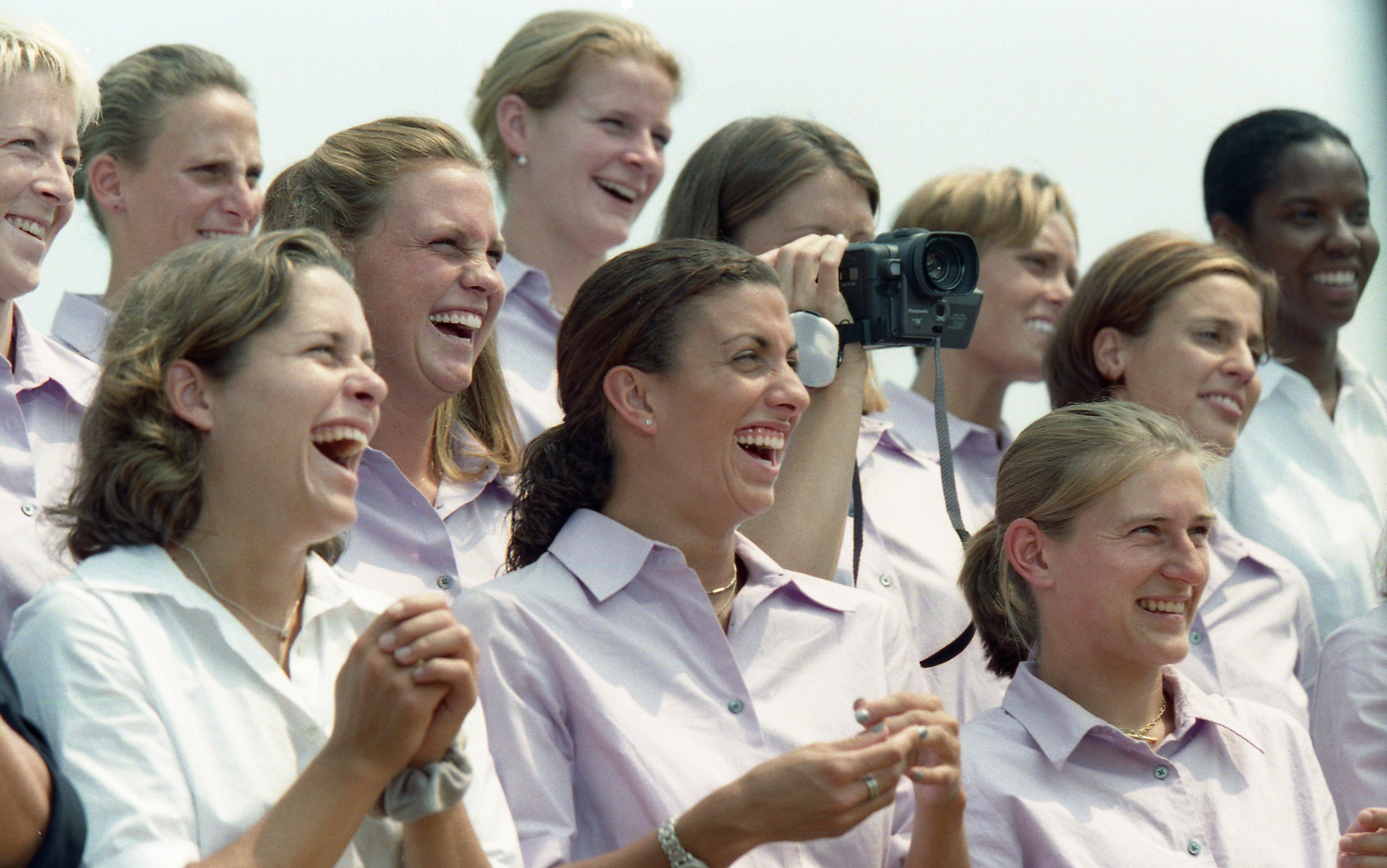 A close-up photograph captures members of a women's soccer team laughing and smiling together outdoors. The women wear matching light purple button-down shirts, except for one player in the front left who wears a white collared shirt. Several players are laughing heartily with wide expressions, while another player in the middle background holds up a small, dark video camera to record. The background features a bright, overcast sky.