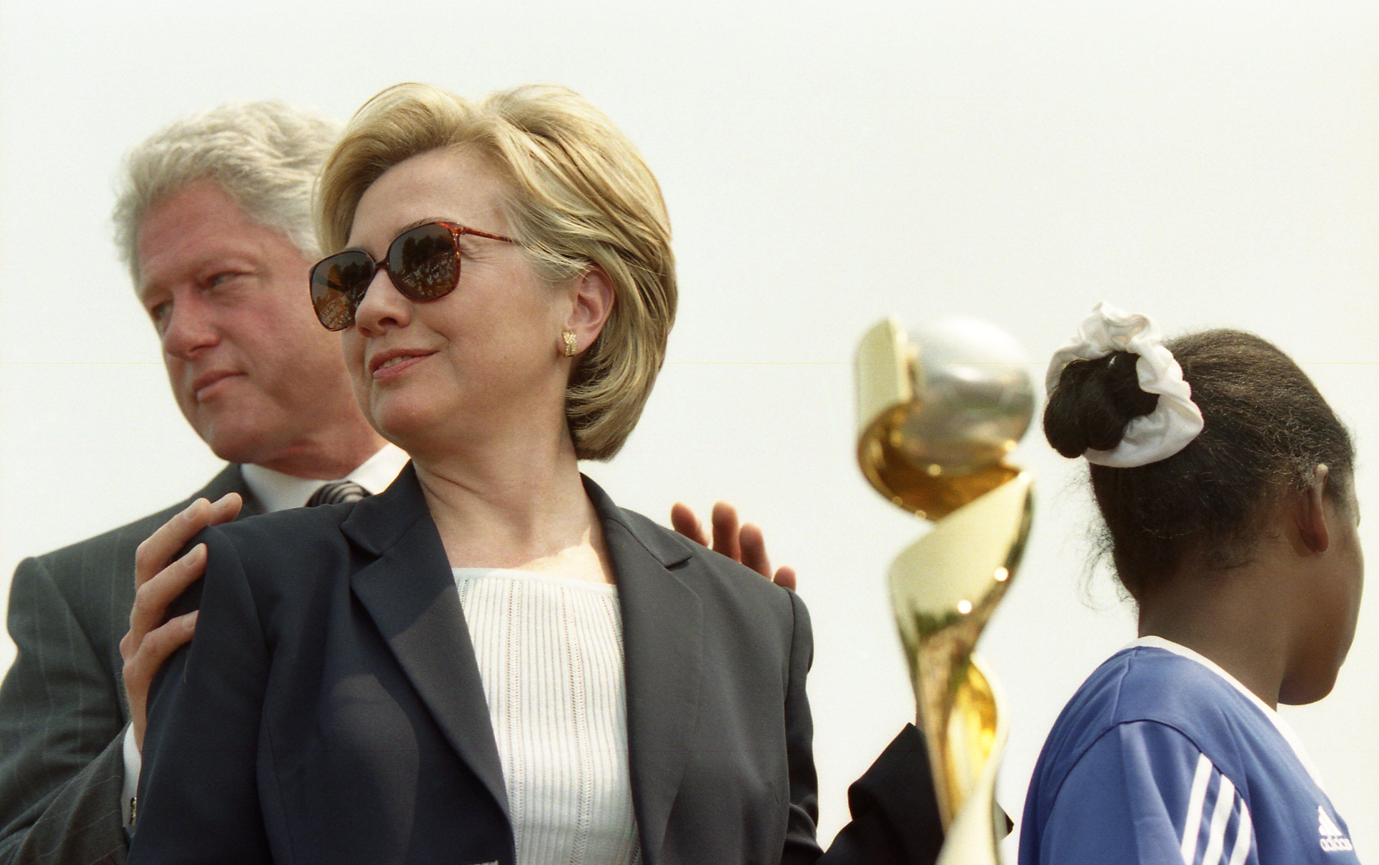 A close-up shot captures First Lady Hillary Clinton looking off-camera while wearing a black blazer, a white top, sunglasses, and gold earrings. Former U.S. President Bill Clinton stands partially behind her, placing his hand on her shoulder. On the right, the back of a young Black girl's head is visible in a blue Adidas sports jersey with a white scrunchie in her hair, alongside a portion of a gold trophy.