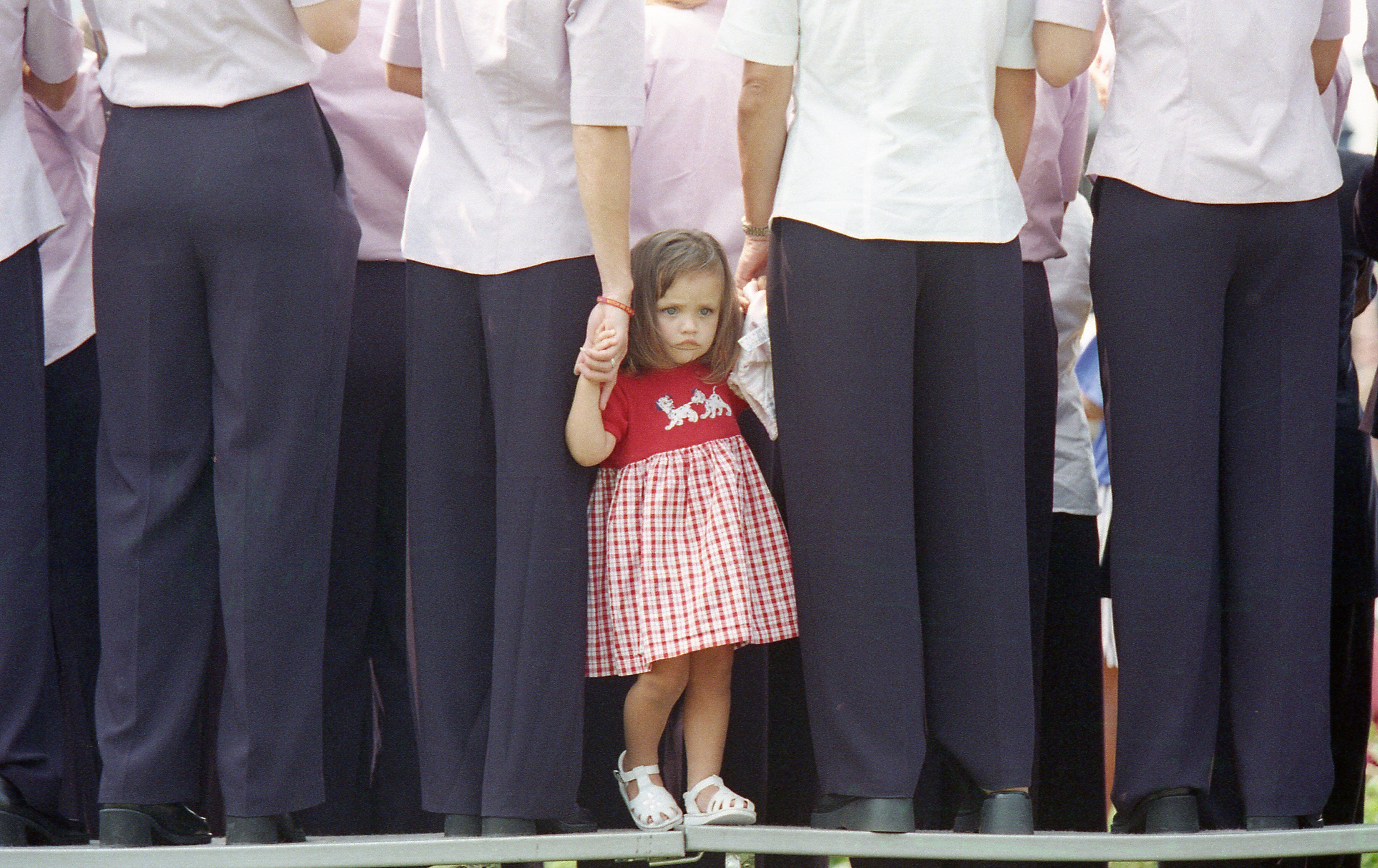 A medium shot from behind focuses on a young girl in a red and white gingham dress standing on a stage, holding the hand of a woman in a light pink shirt and dark pants. The girl looks toward the camera with a serious expression, framed by the legs and backs of multiple adults standing in line wearing matching pink or white shirts and dark trousers.