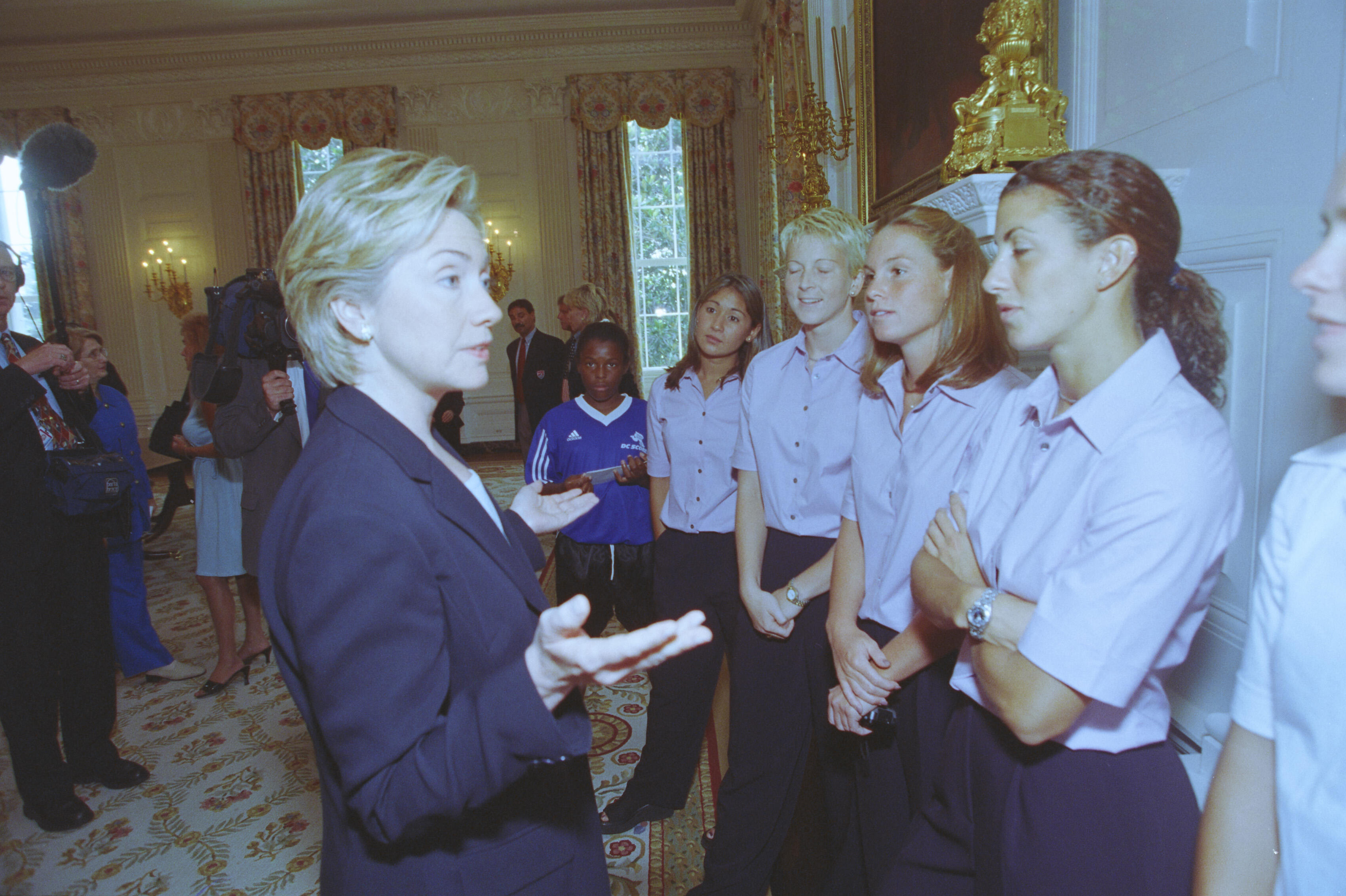 A medium shot captures First Lady Hillary Clinton from a side profile as she gestures with open hands while speaking to a group of listening women. The women stand closely together in a line, wearing matching light pink short-sleeved button-down shirts and dark pants. In the background, a young girl in a blue sports jersey stands nearby, while multiple news cameras and media personnel film the gathering in a brightly lit room decorated with floral curtains and an ornate gold fireplace mantel.